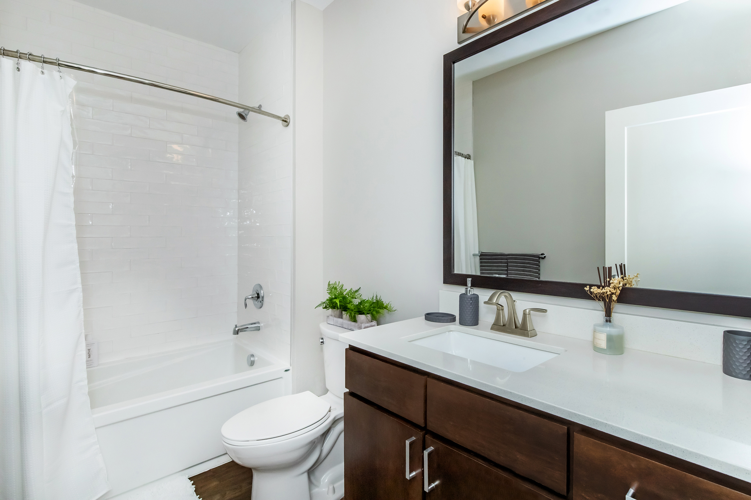 A modern bathroom featuring a white bathtub with a shower curtain, a dark wood vanity with a sink, and a large decorative mirror. There are plants and accessories on the vanity, and light-colored walls create a bright, clean atmosphere.