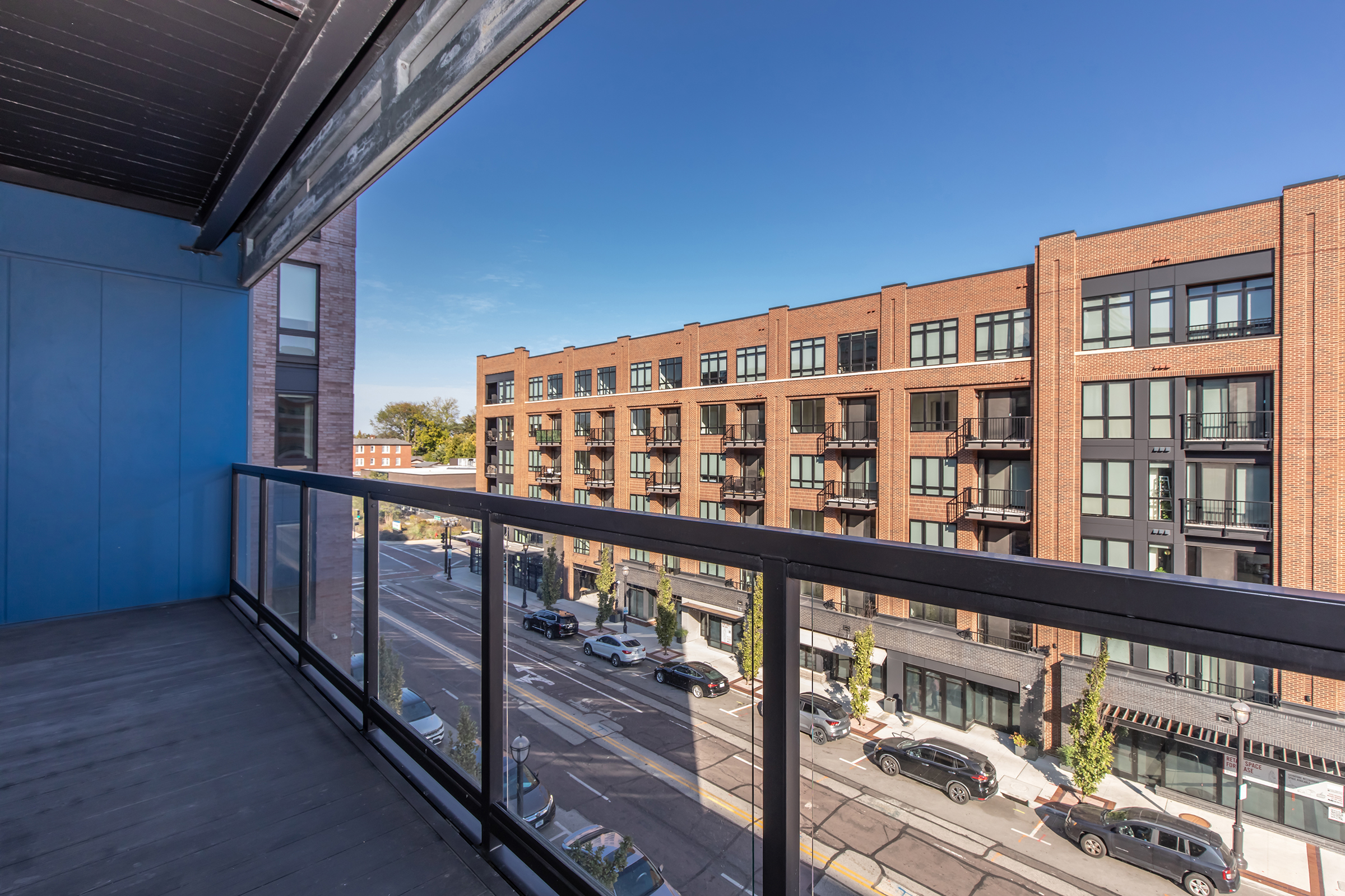A view from a balcony overlooking a bustling street. The balcony features a modern design with glass railings, while the street below is lined with parked cars and brick buildings with balconies. The sky is clear and blue, adding to the urban atmosphere.
