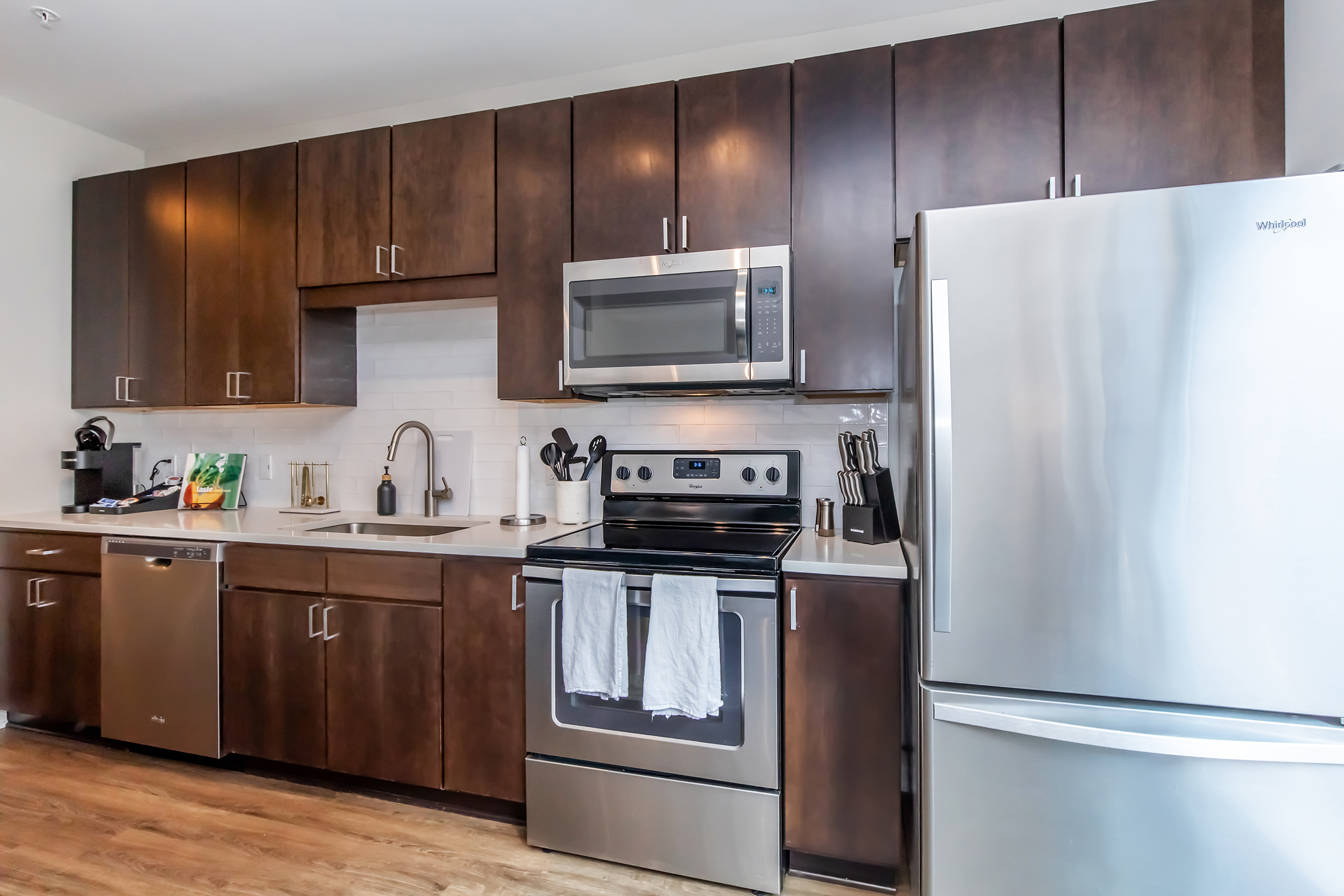 Modern kitchen featuring dark wood cabinetry, stainless steel appliances including a refrigerator, microwave, and oven. The countertop is light-colored with a sink and kitchen utensils visible. A coffee maker is placed on the countertop, and the overall decor is sleek and contemporary.