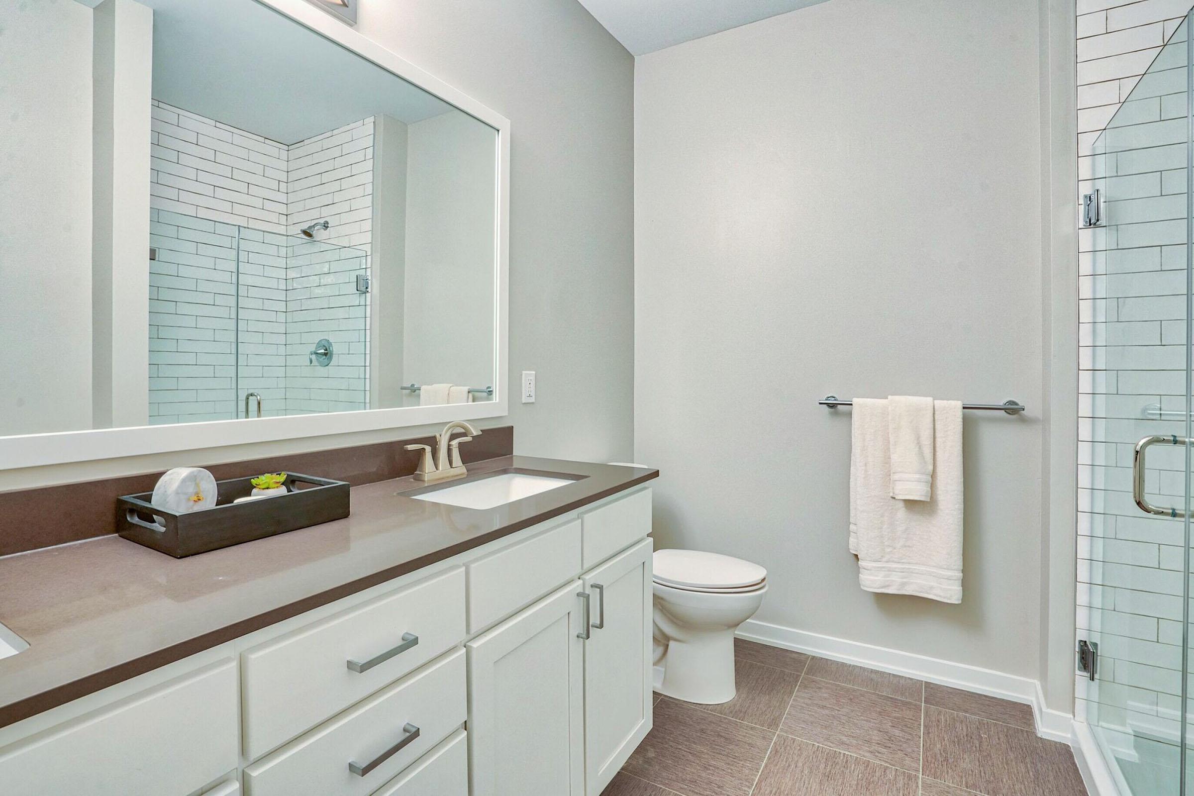 A modern bathroom featuring a sleek design with a large mirror above a double sink. The space includes a glass shower with white tiles, a toilet, and a towel hanging on a rack. The countertop is dark brown, and there is a decorative tray on the sink. Bright, neutral walls give a clean, open feel.