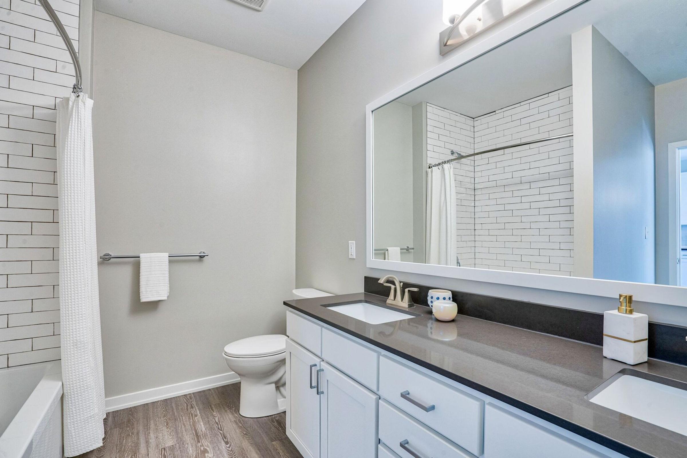 A modern bathroom featuring a white bathtub with a gray shower curtain, a large mirror above a dark countertop with dual sinks, and stylish fixtures. The walls are painted a light color, and a towel hangs on a bar. The floor is a wood-like material, giving the space a contemporary feel.