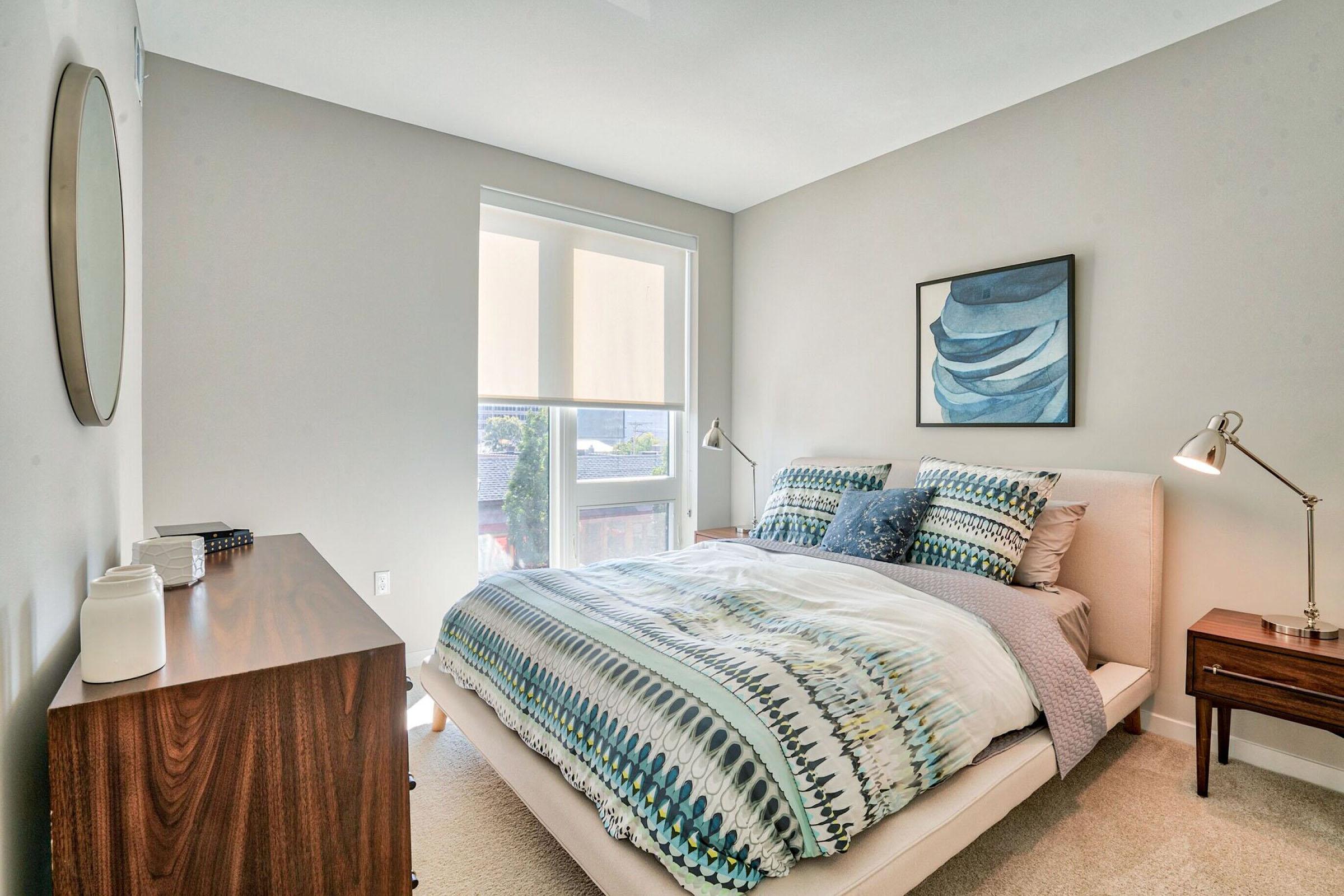 Cozy bedroom featuring a large bed with a patterned duvet, two bedside lamps, and a piece of artwork on the wall. The room is well-lit by natural light coming through a window, and there's a modern dresser made of wood, adding to the contemporary decor.
