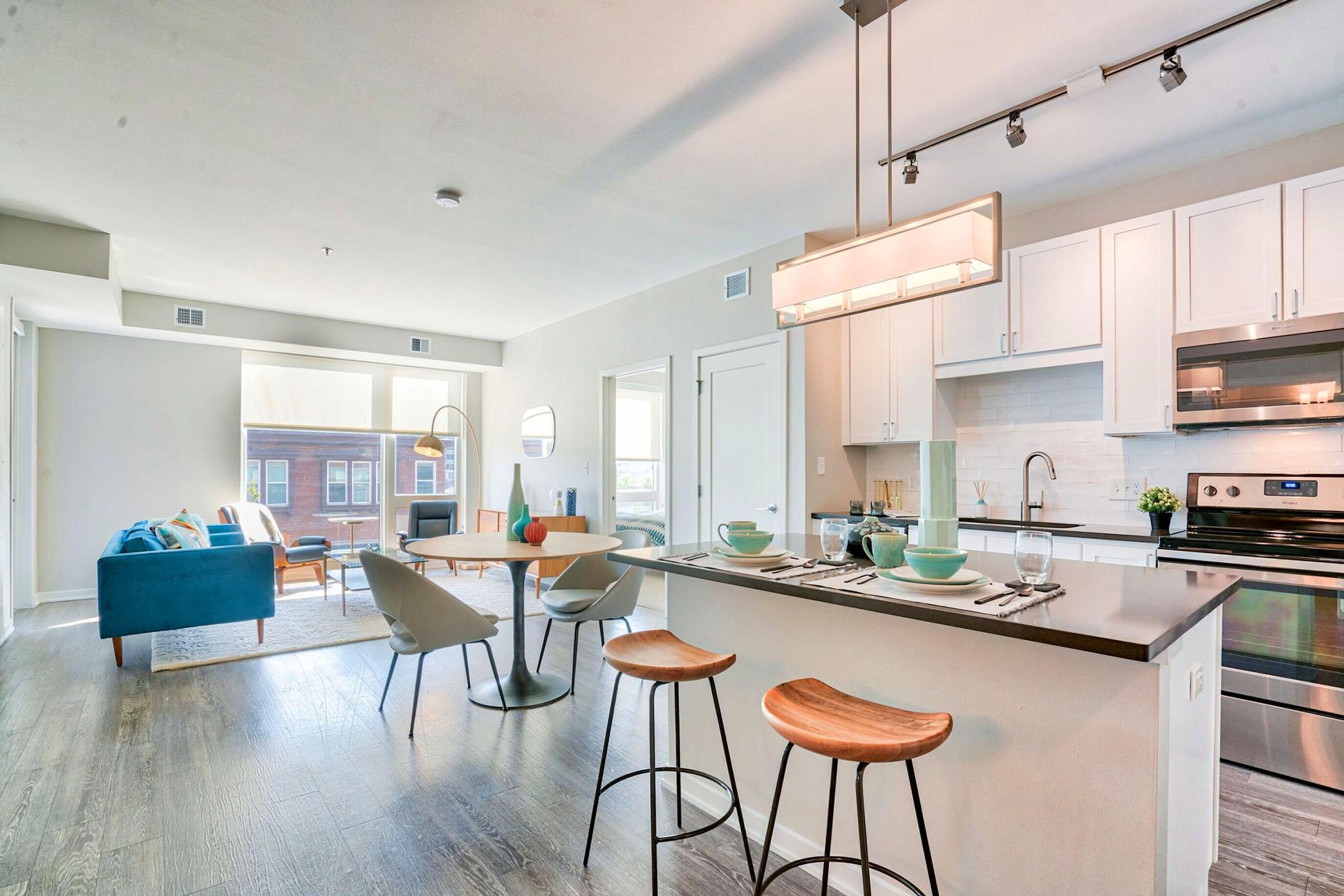 Modern kitchen and living area in an open-concept apartment. The kitchen features stylish cabinetry, stainless steel appliances, and a countertop with seating. A cozy living area includes a blue couch, a coffee table, and natural light streaming through large windows, creating a bright and inviting atmosphere.