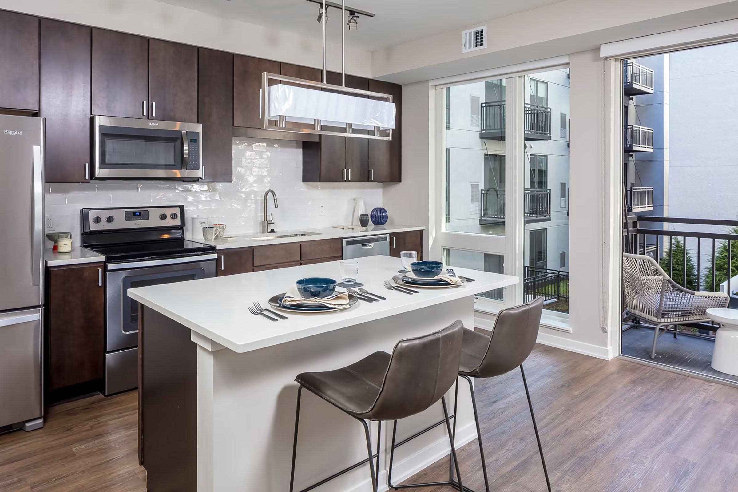Modern kitchen featuring dark wood cabinetry, stainless steel appliances, and a white countertop. An island with seating for two is set with plates and cutlery. Large windows let in natural light, revealing a balcony and an exterior view of a multi-story building.