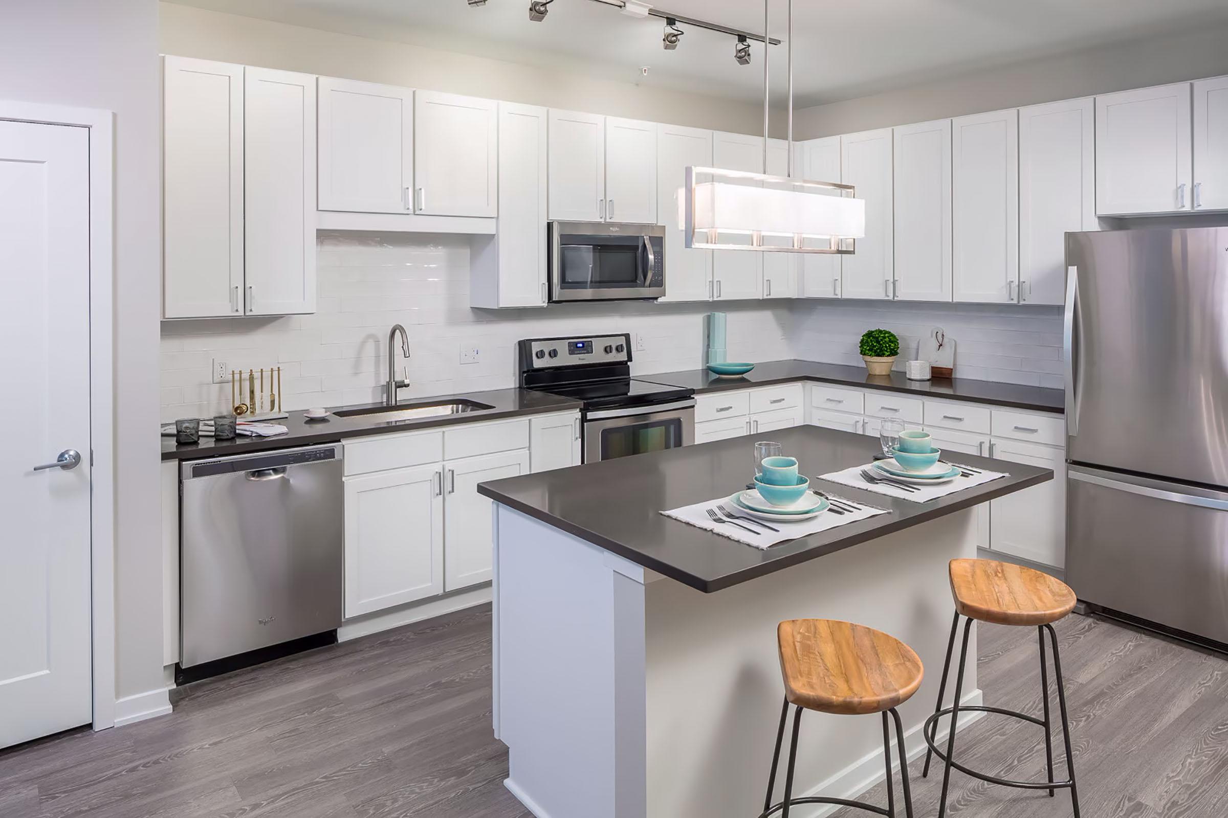 Modern kitchen featuring white cabinetry, stainless steel appliances, and a large island with two wooden bar stools. The countertop is dark, contrasting with the light cabinets. A stylish light fixture hangs above the island, and there are place settings with teal dishware on the counter. The overall decor is contemporary and minimalist.