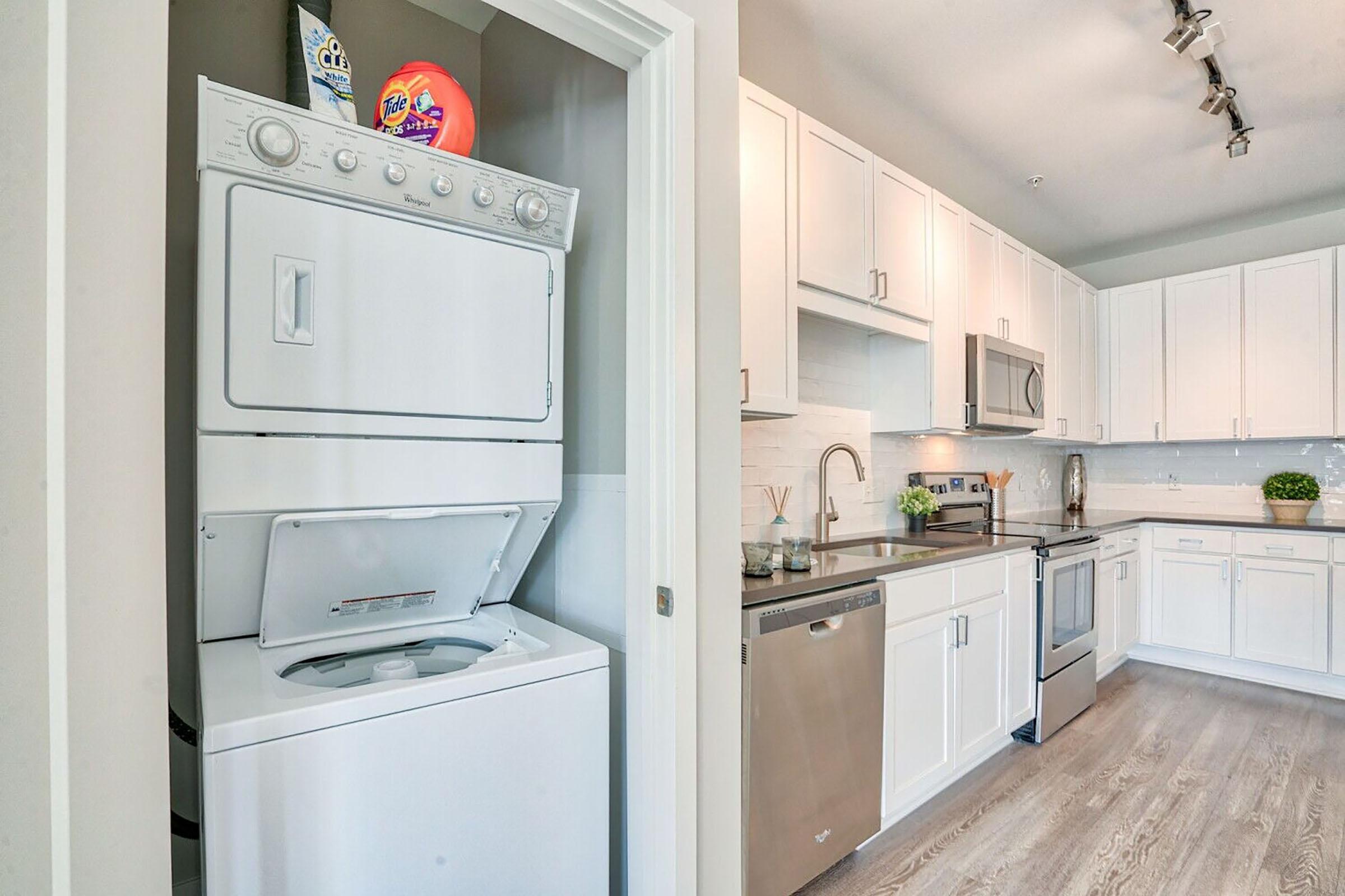 A modern kitchen featuring white cabinetry, stainless steel appliances, and a stylish backsplash. On the left, there is a stacked washer and dryer, while the kitchen area showcases a countertop with a sink and decorative plants. Natural light fills the space, enhancing its contemporary feel.