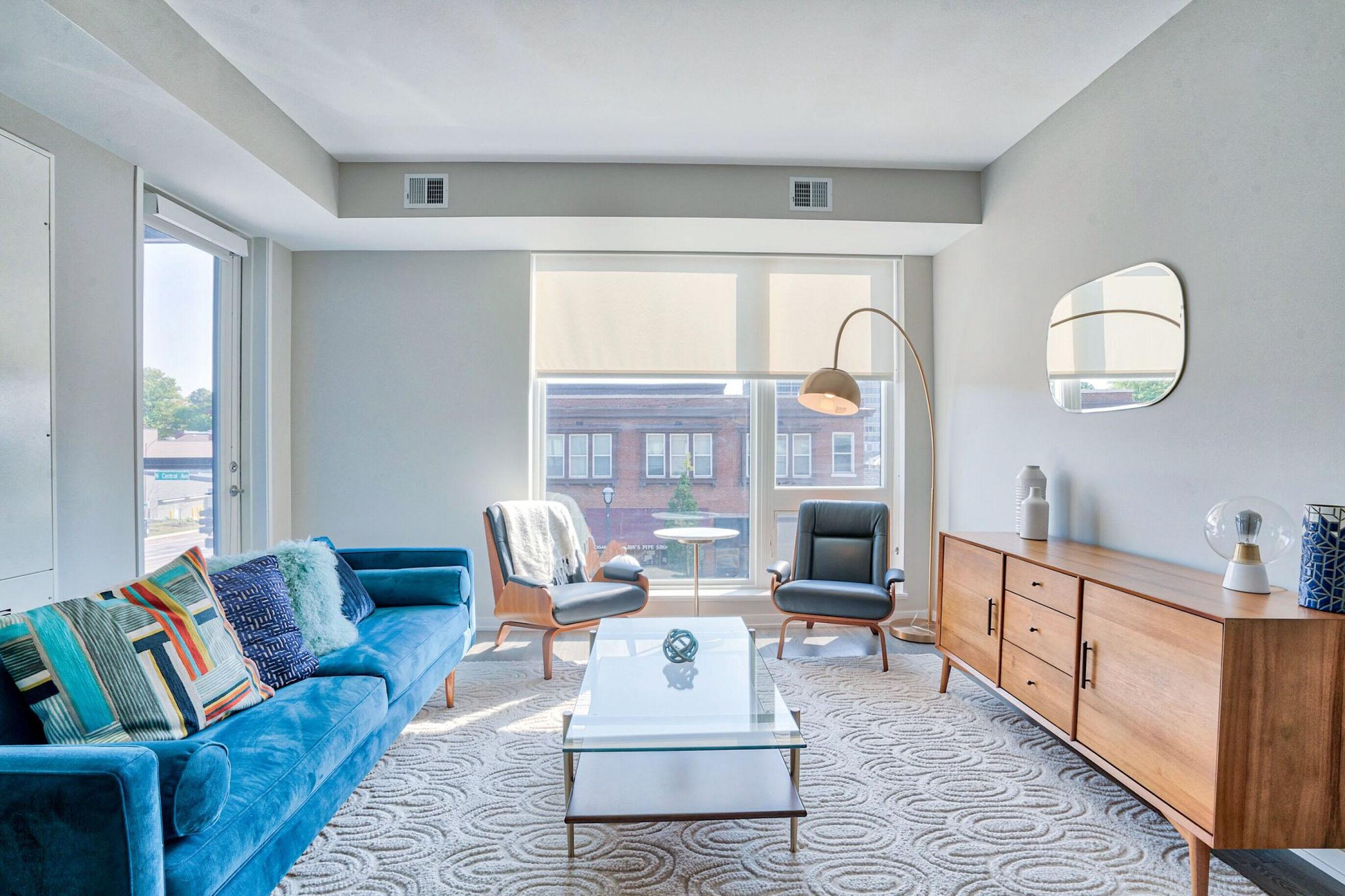 A modern living room featuring a blue sofa with patterned cushions, a glass coffee table, and a wooden sideboard. Two stylish chairs are positioned near a large window that allows natural light in, revealing an urban view outside. Minimalist decor elements, including a floor lamp and decorative objects, add a contemporary touch.