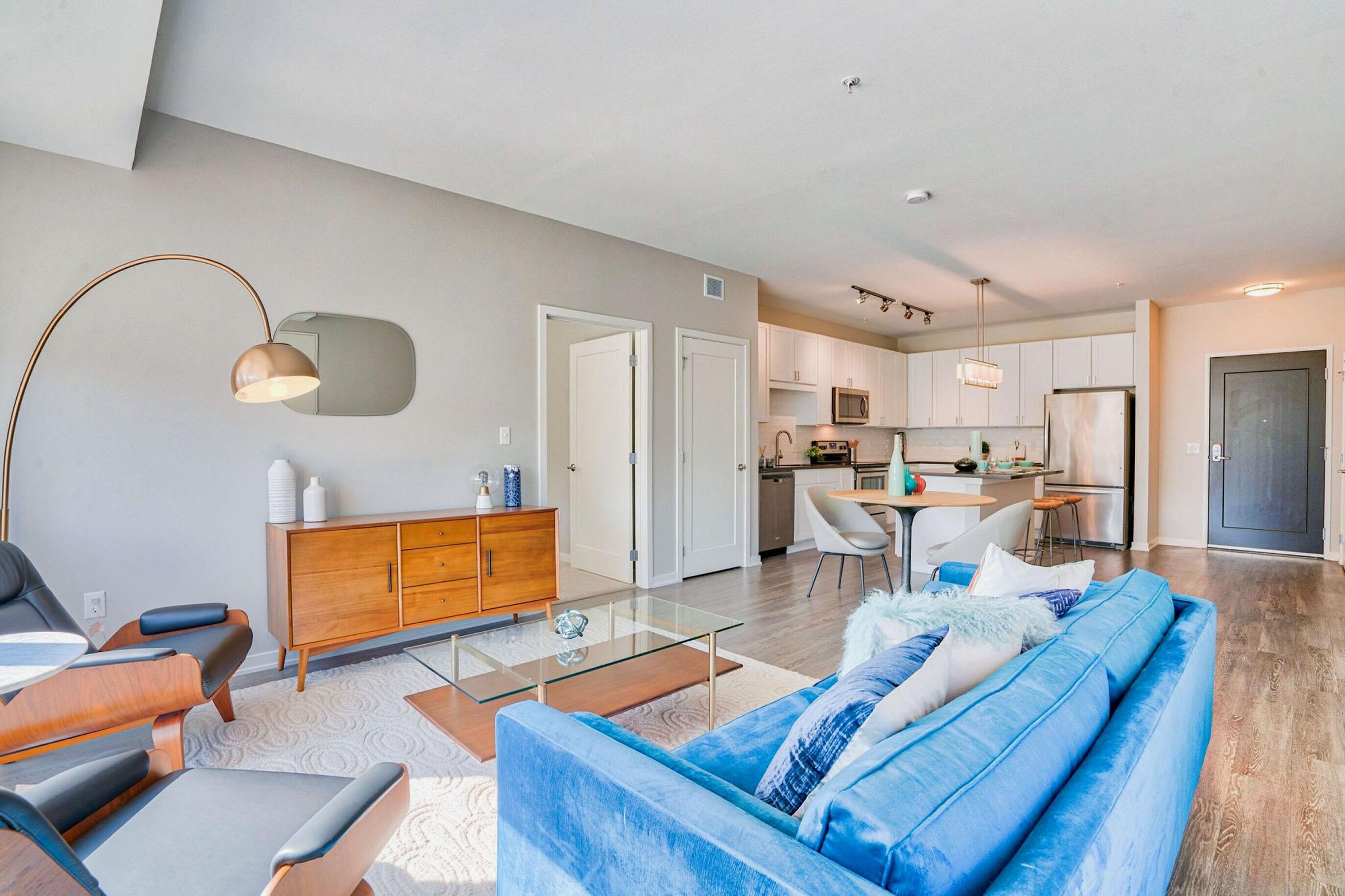 A modern living space featuring a blue sofa, wooden furniture, and a sleek coffee table. The room has large windows allowing natural light, with a dining area visible in the background. Neutral-colored walls and contemporary decor create a stylish and inviting atmosphere.