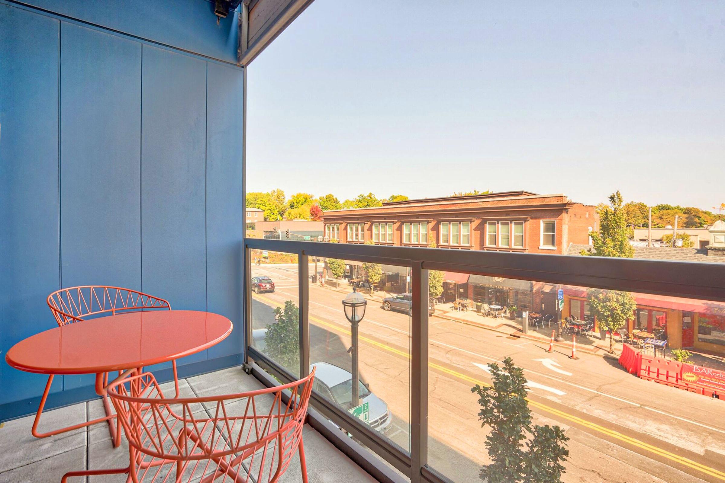 A balcony view featuring orange wire furniture, including a table and two chairs. The railing provides a clear view of a street with shops and trees, under a clear blue sky. The atmosphere suggests a lively urban environment.