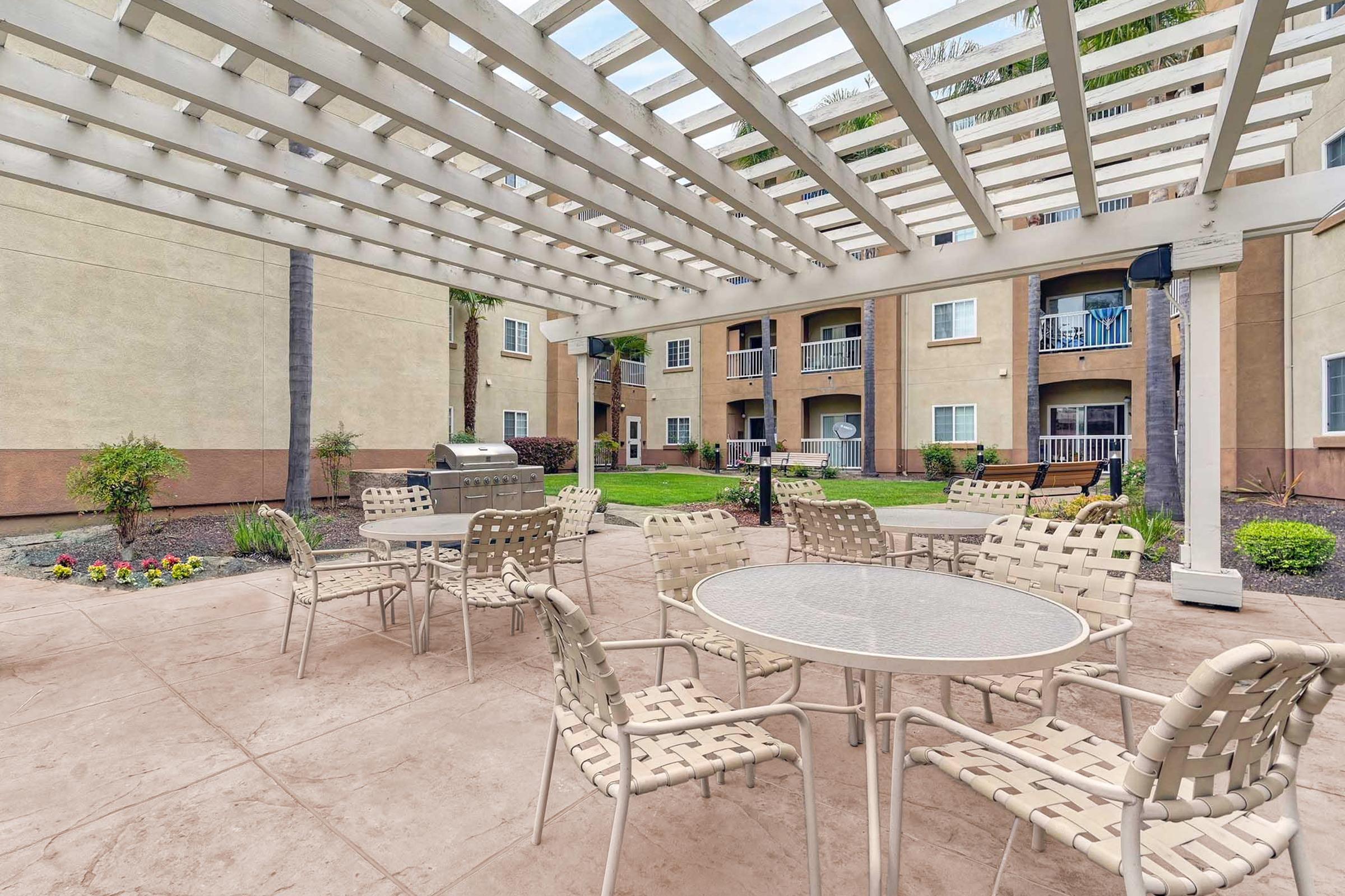 Outdoor lounge area featuring circular tables and chairs under a white trellis. In the background, an apartment building is visible with balconies and green landscaping. A barbecue grill is located nearby, creating a welcoming space for relaxation and social gatherings.