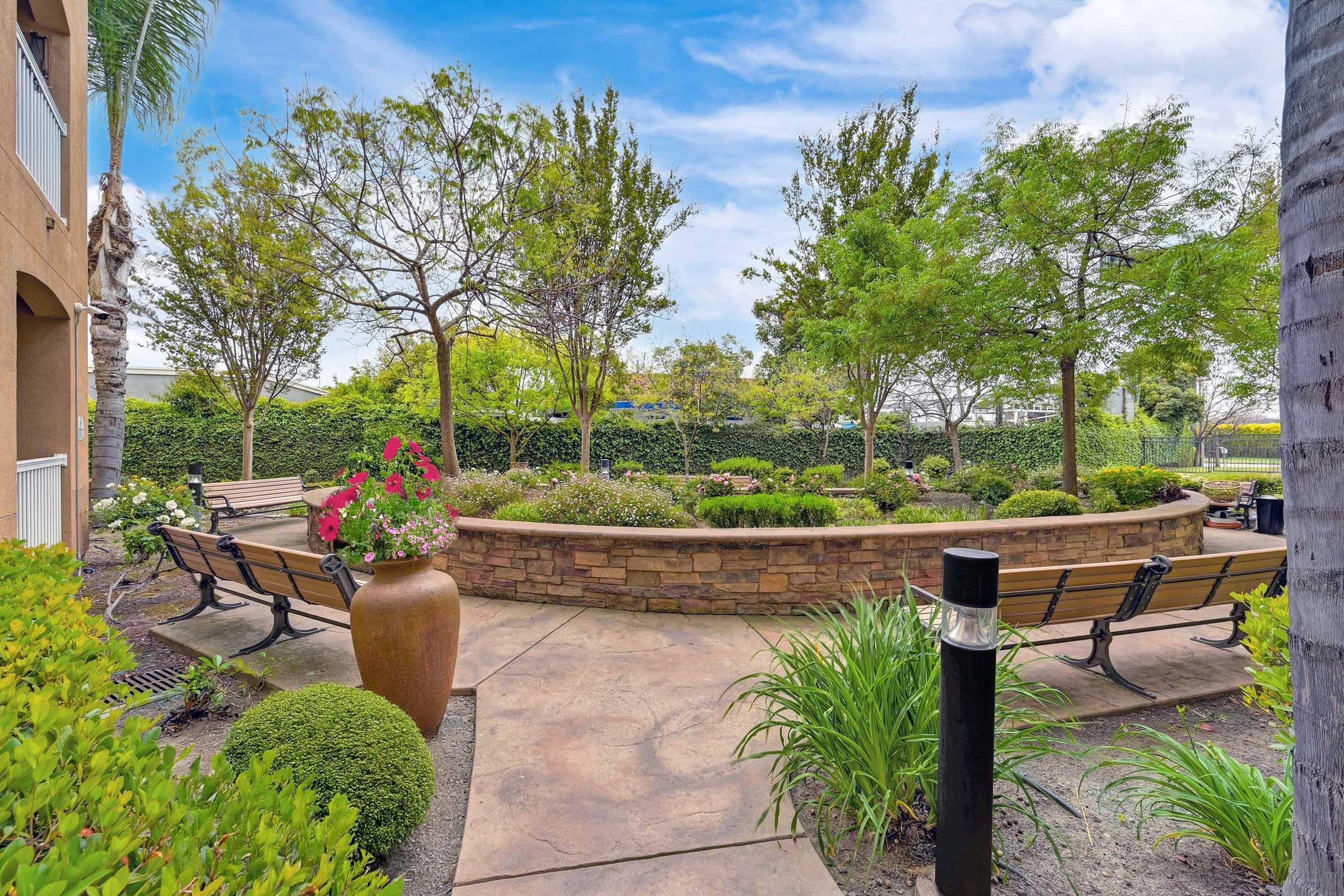 A landscaped outdoor area featuring a stone flower bed surrounded by benches, vibrant flowers, lush green plants, and trees under a blue sky. The peaceful setting invites relaxation and enjoyment of nature.