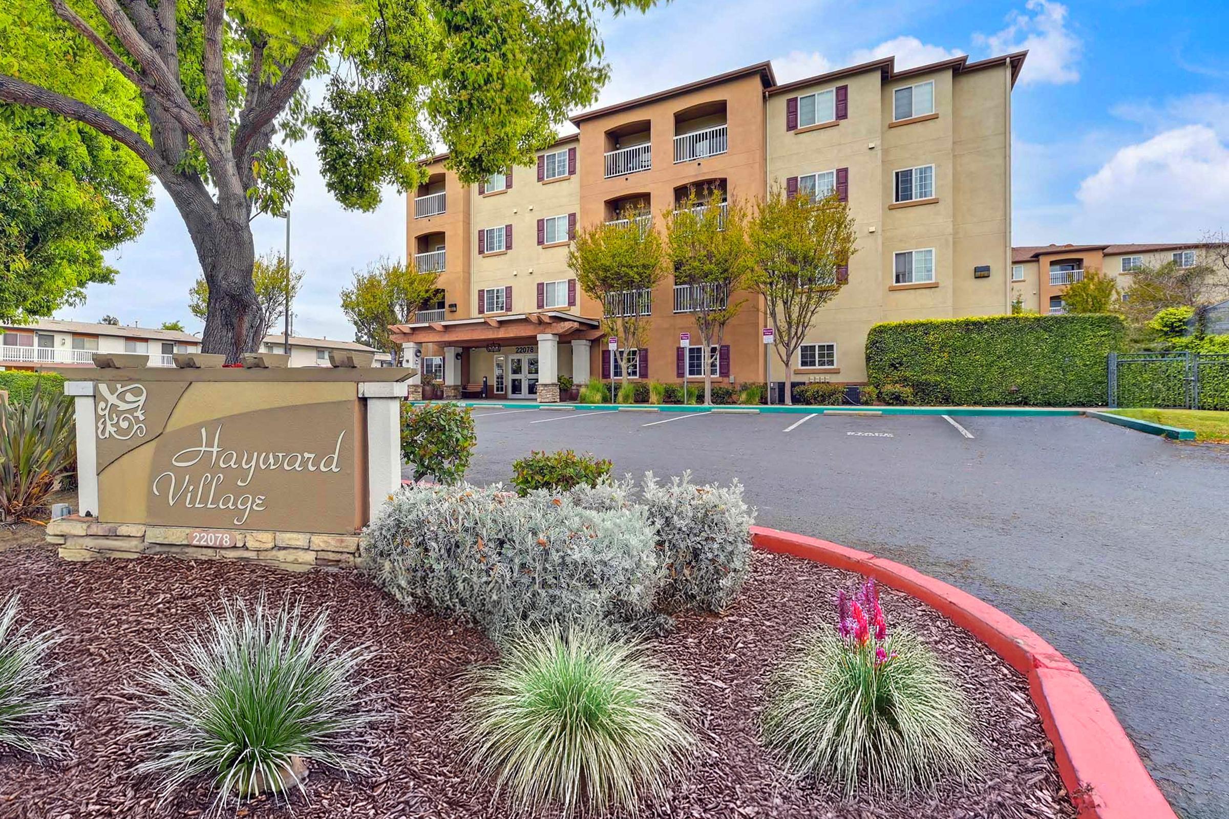 A multi-story residential building with balconies and windows, surrounded by greenery. In the foreground, there is a sign for "Hayward Village" along with landscaped shrubs and flowers. The parking area is visible, and the sky is partly cloudy.