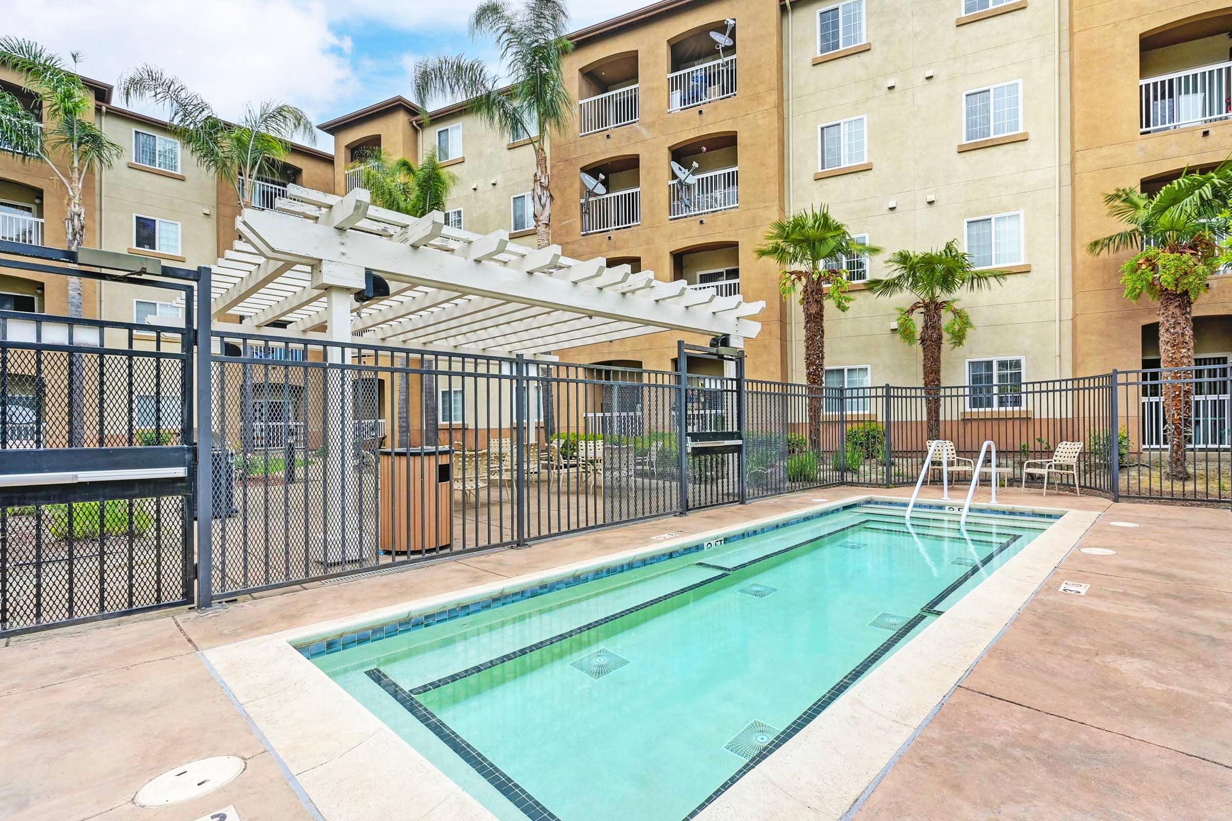 A small, clean swimming pool surrounded by a gated area, with a pergola overhead. Palm trees and greenery enhance the tropical atmosphere, and an apartment building with multiple balconies is visible in the background. The sky is partly cloudy, adding to the pleasant outdoor setting.