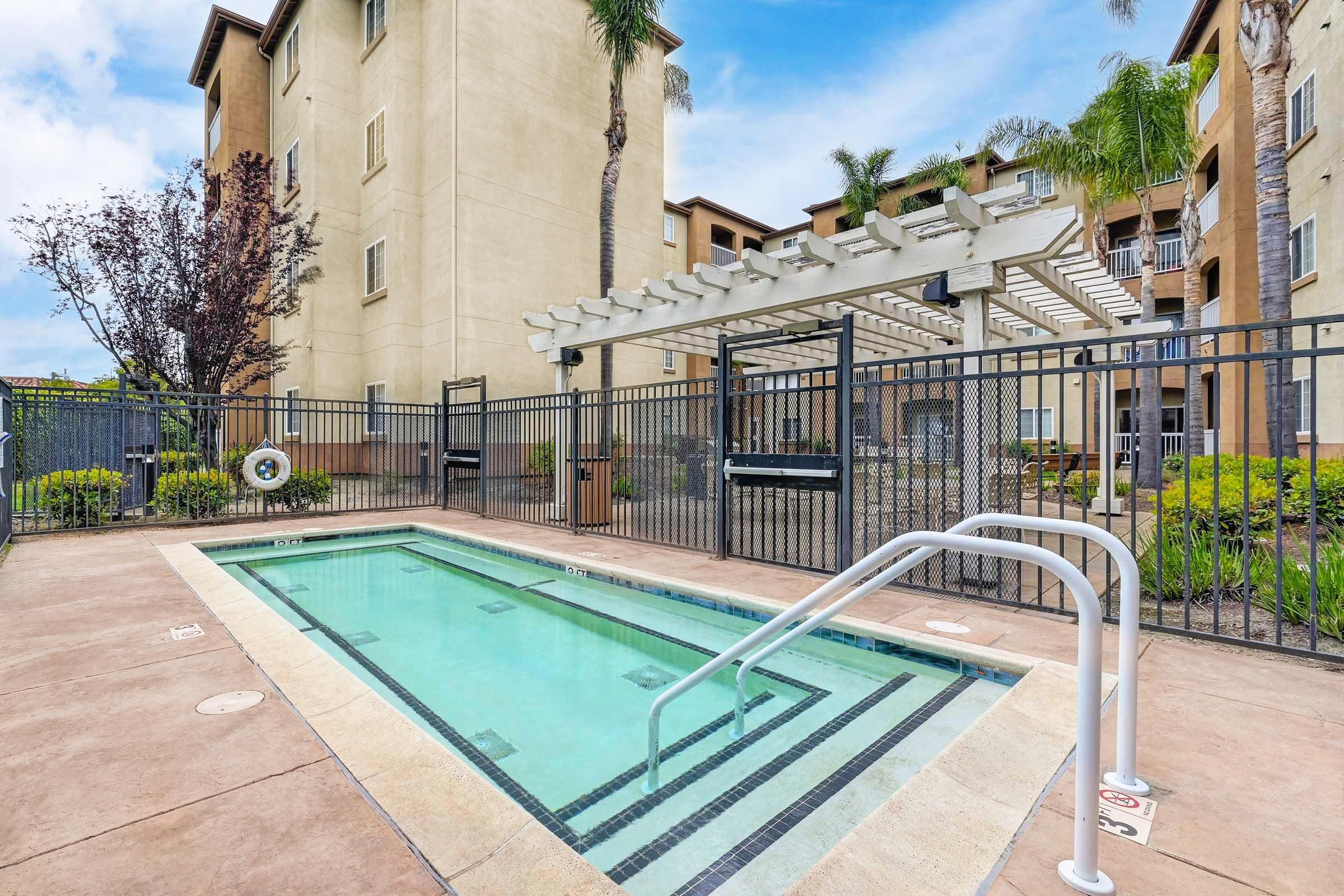 A small outdoor swimming pool with a shallow area, surrounded by a gated fence and featuring a covered seating area. Lush greenery and palm trees are visible nearby, with an apartment building in the background under a partly cloudy sky.