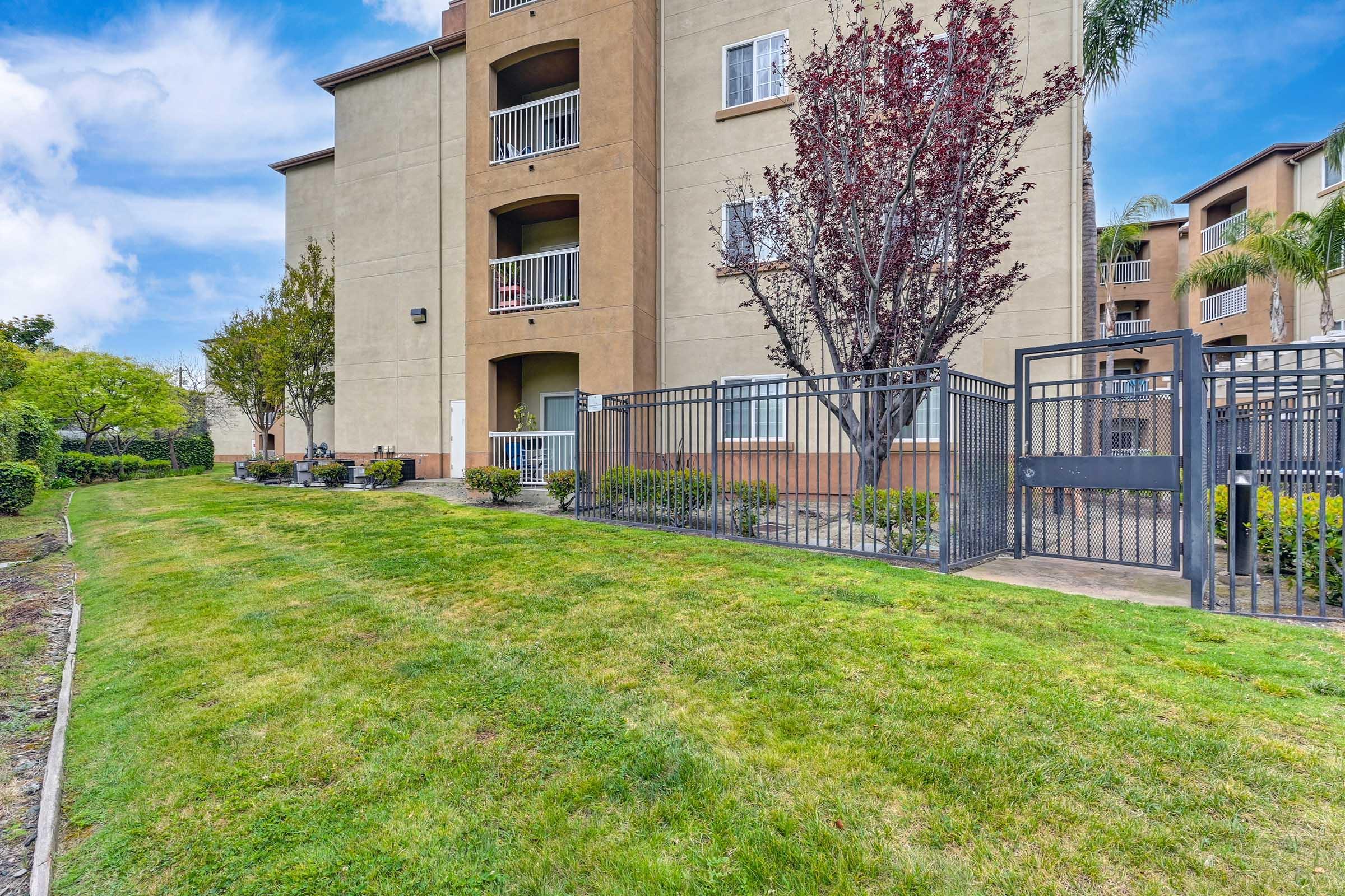 A well-maintained apartment building with a light brown exterior, featuring balconies. In front, there is a green lawn bordered by decorative plants and trees. A black gate leads to a fenced area, enhancing the property's security and aesthetics. The sky is clear with a few clouds.