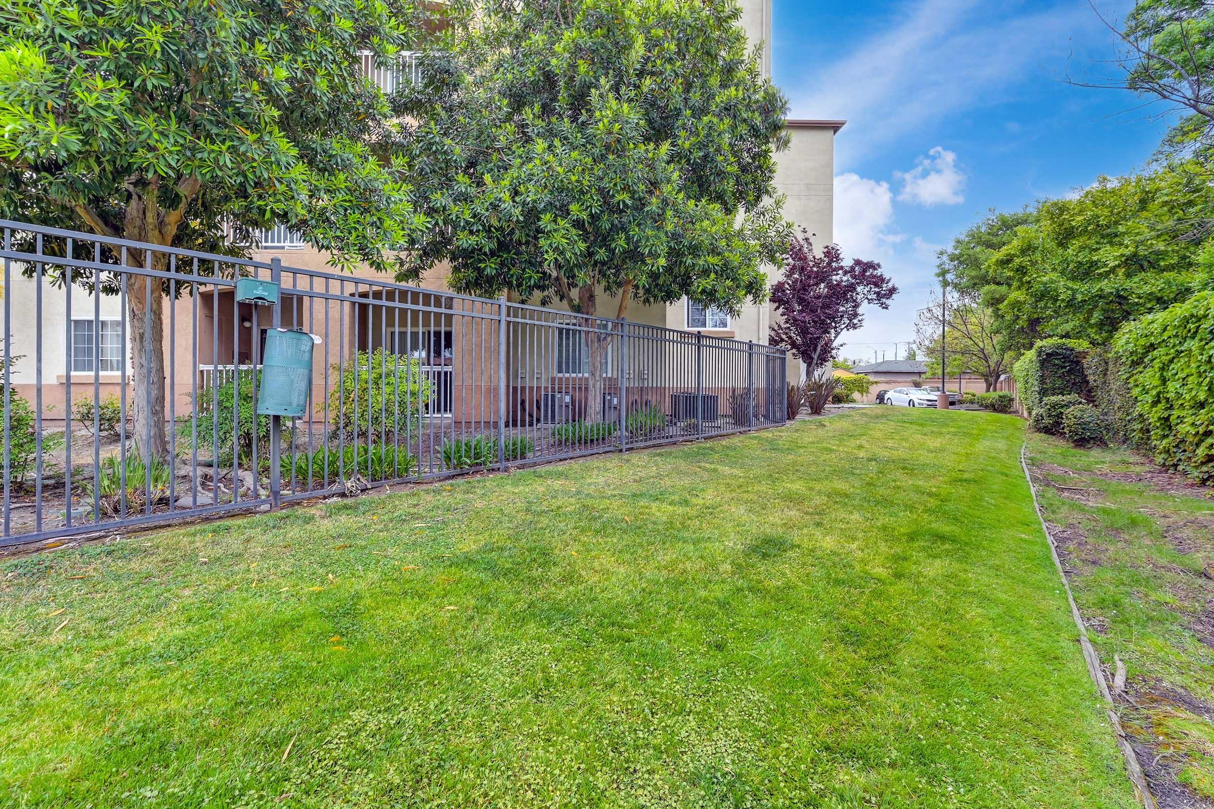 A grassy area lined with greenery, featuring a fence on one side. Nearby, there are shrubs and small trees, along with a building in the background. The sky is clear with a few clouds, creating a bright and inviting atmosphere. The scene appears well-maintained and tranquil.