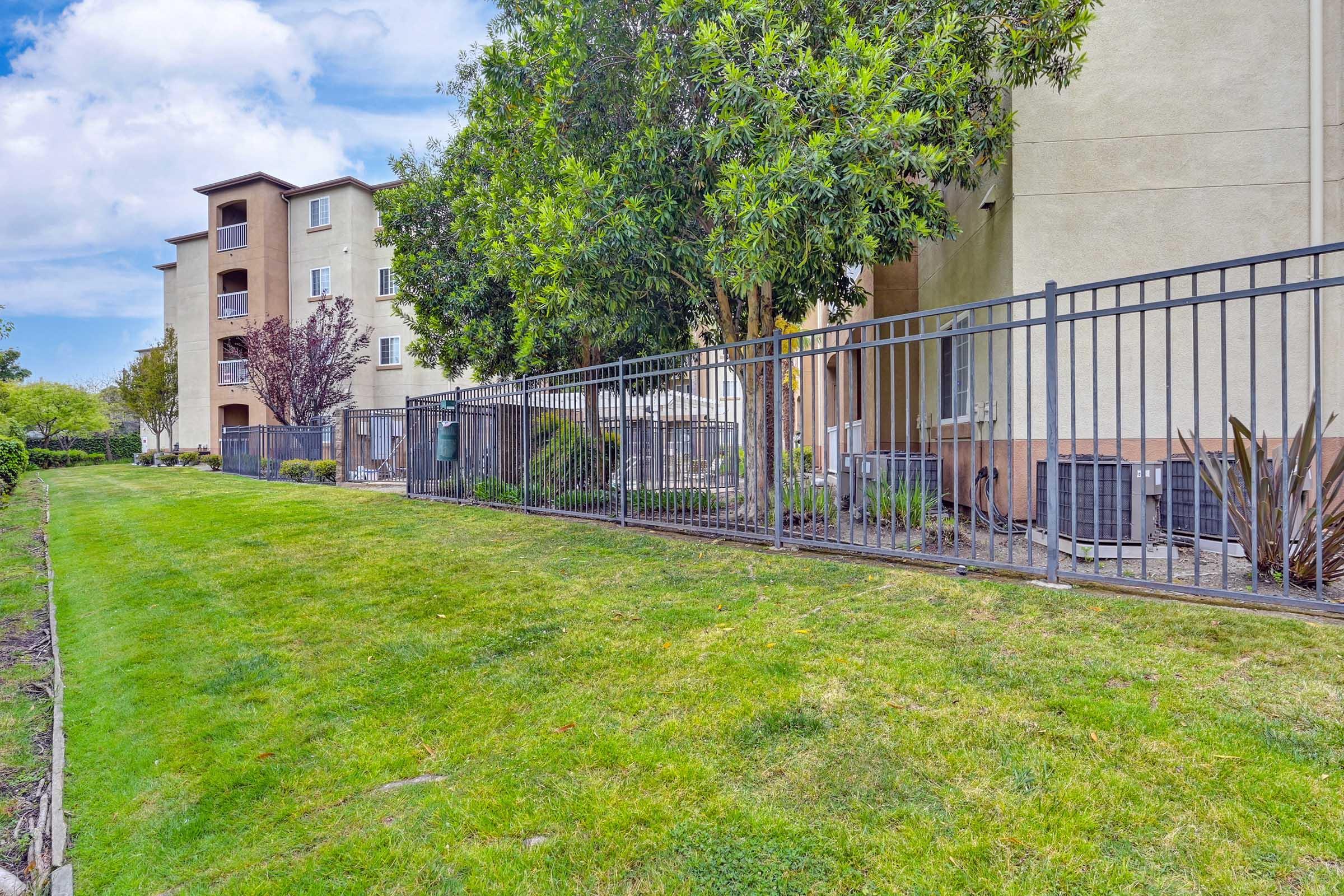 A landscaped area with green grass in front of a multi-story residential building. A fence borders the property, with several air conditioning units visible along the building's side. Trees and shrubs add to the greenery, and a partly cloudy sky is overhead.