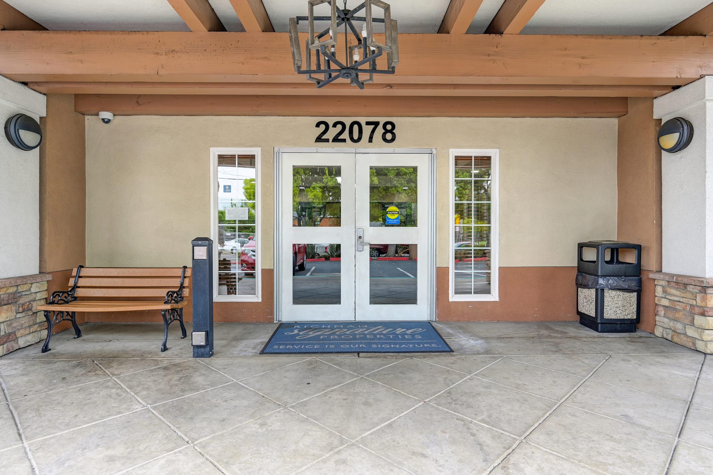 Entrance of a building featuring large glass double doors with the number 22078 above. There is a bench nearby and a trash can on the right. The area is paved with stone tiles, and there are trees visible in the background. A doormat with a welcome message is placed in front of the doors.