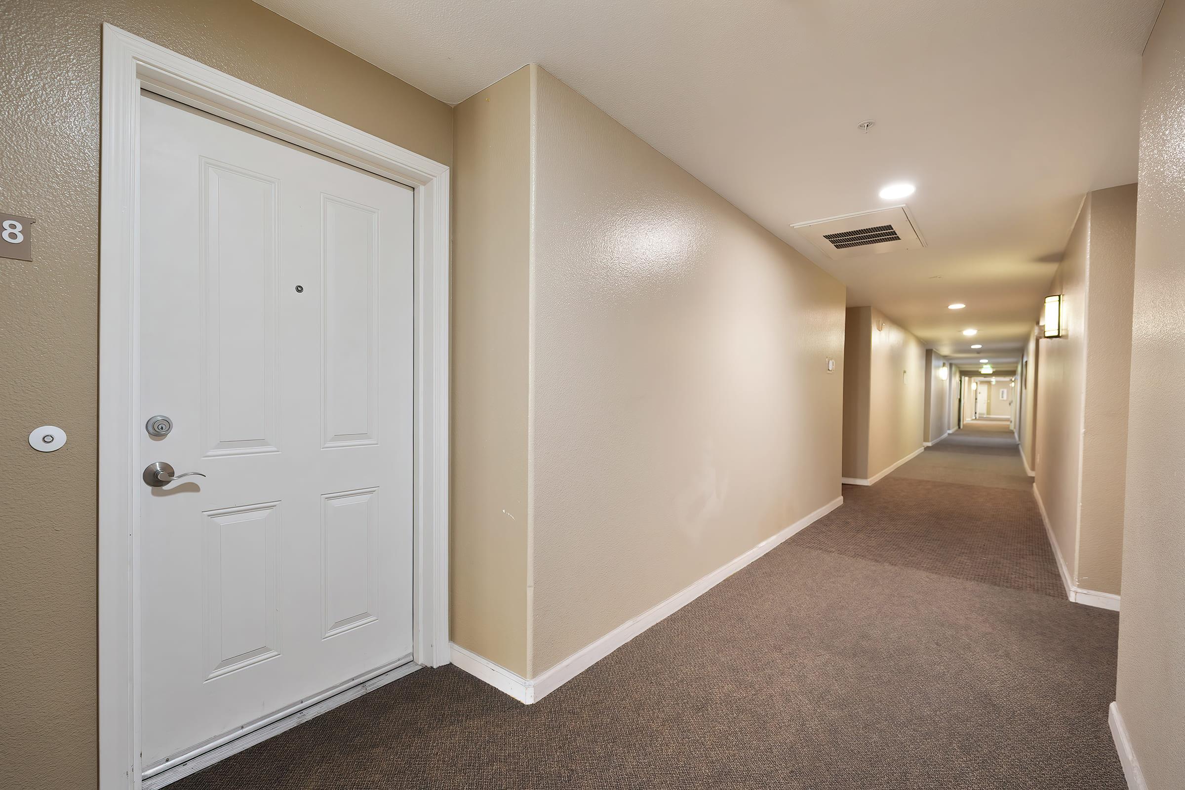 A view of a hotel corridor featuring a light-colored wall and carpeting. A white door is visible on the left, labeled with the number 8. The well-lit hallway extends into the distance with additional doors and soft lighting along the walls, creating a clean and inviting atmosphere.