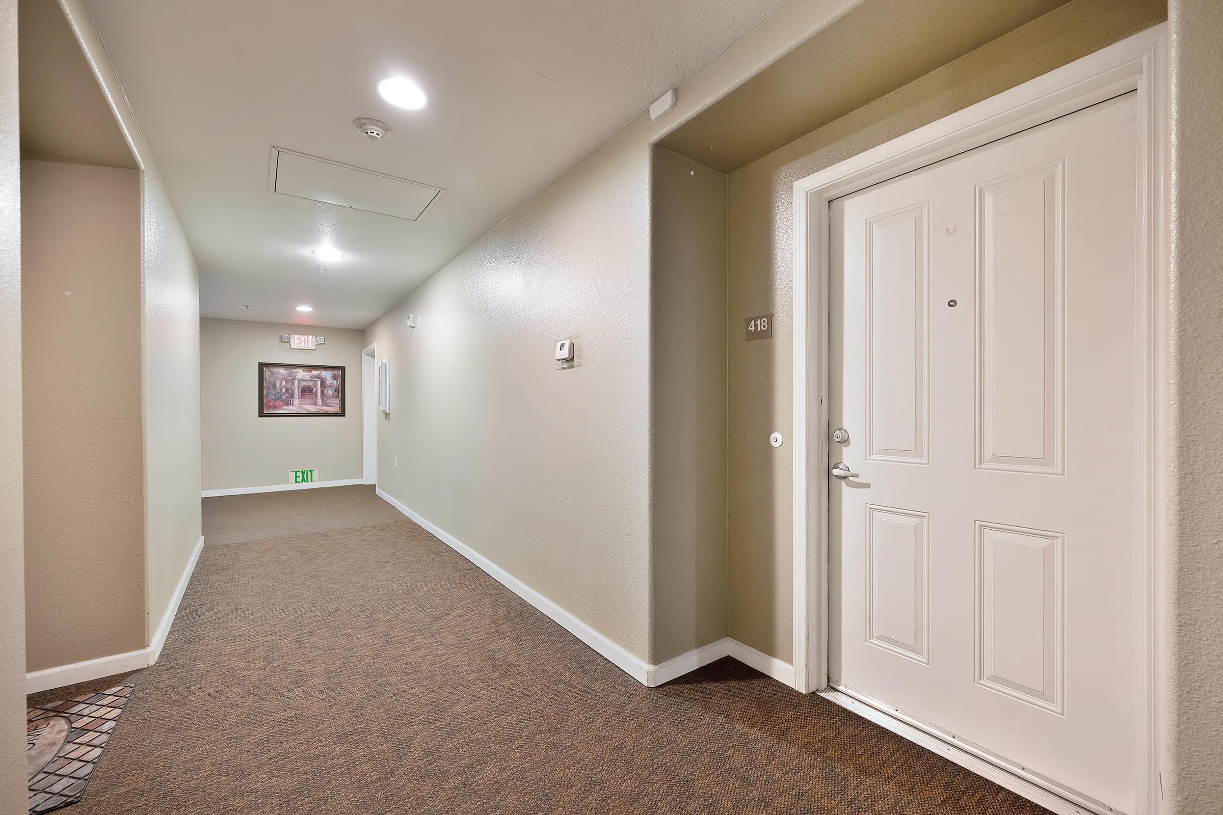 A well-lit hallway featuring beige walls and carpeted floors. A closed door labeled "418" is visible on the right, with a small decorative entry mat in front. The hallway has wall-mounted light fixtures and a few exit signs in the background, creating a clean and inviting atmosphere.