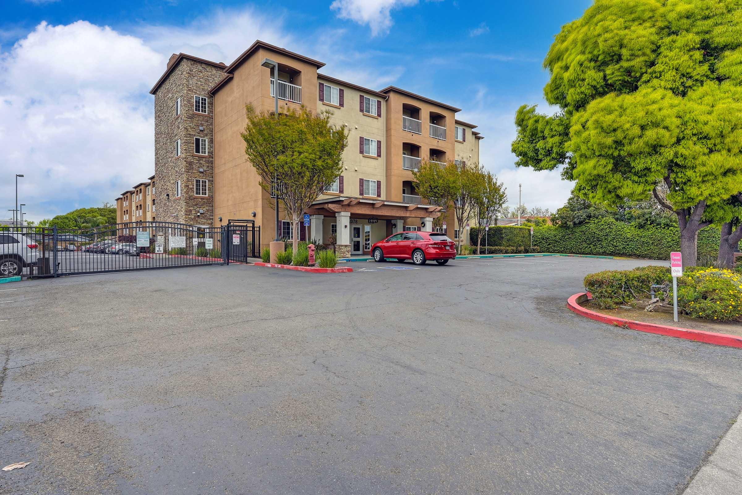 A three-story building with a stucco facade and stone accents is seen from a parking lot. A red car is parked near the entrance, and the area is lined with small trees and green shrubbery. The sky is partly cloudy, creating a pleasant atmosphere. A gated entrance is visible in the foreground.