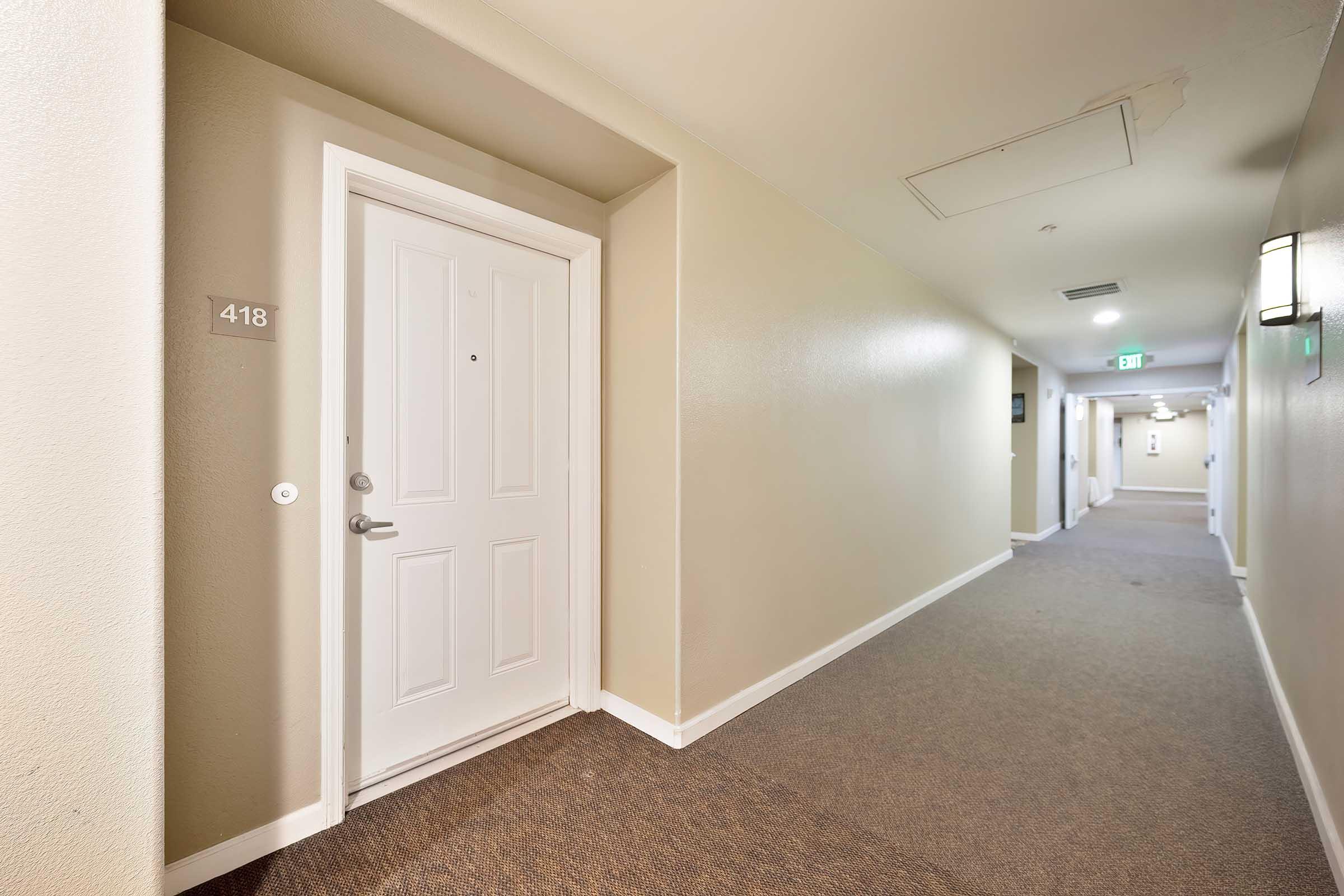 A well-lit hotel hallway with beige walls and carpet. A closed door labeled "418" is on the left side, with a visible doorbell. The corridor extends towards more doors in the background, with soft overhead lighting and a modern, clean appearance.
