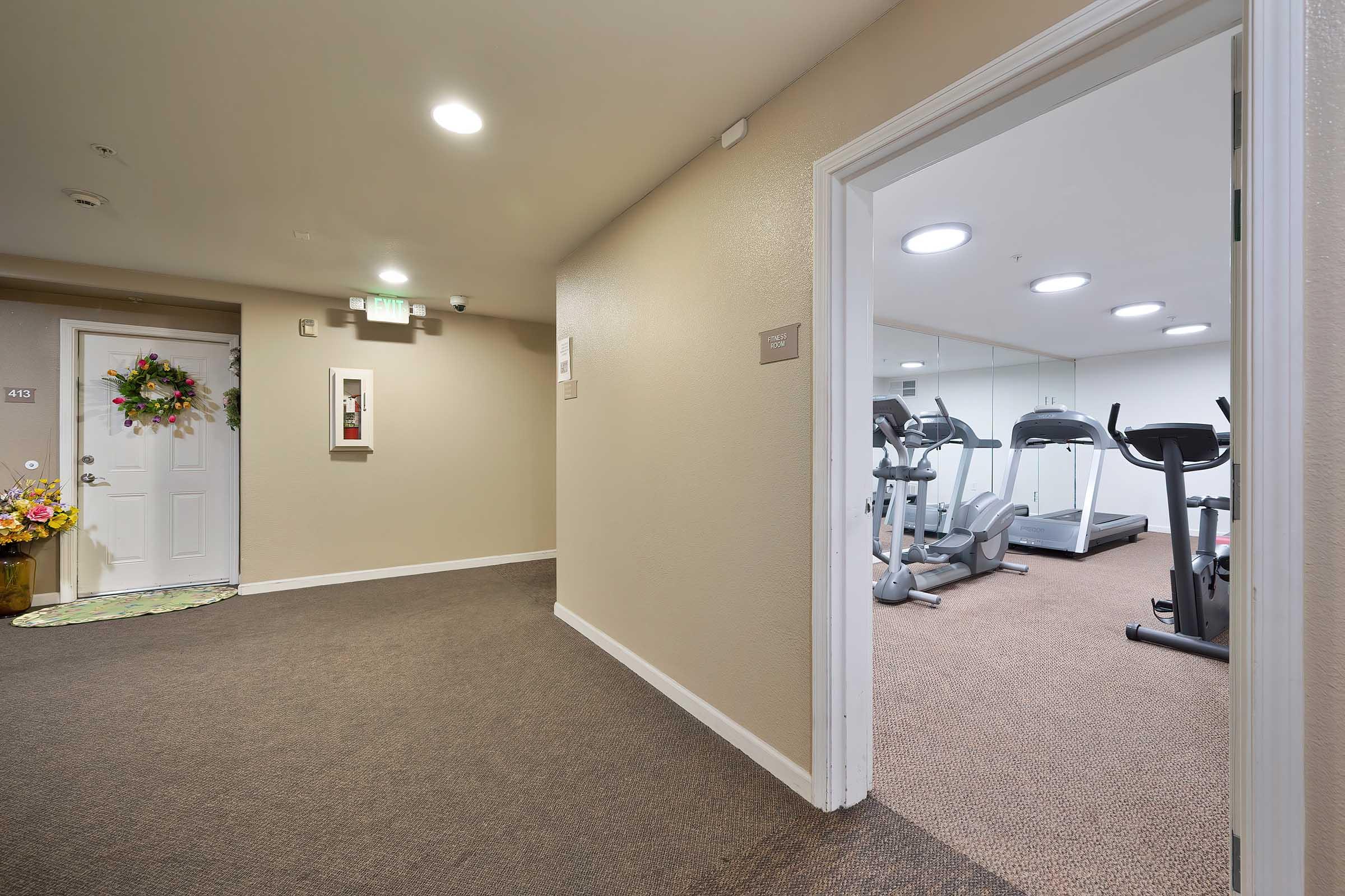 A hallway leading to a fitness room. The corridor features a door with a wreath and a fire extinguisher on the wall. The fitness room is visible through an open door, showcasing treadmills and a mirrored wall. The flooring is carpeted, and the walls are painted in neutral tones.