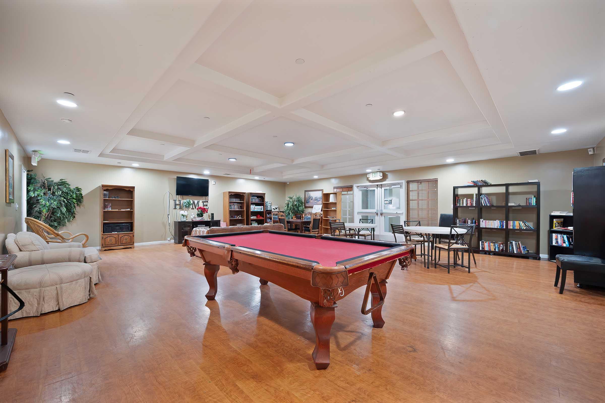 A spacious indoor recreational area featuring a billiard table, comfortable seating, bookshelves, and a dining table. The room is well-lit with recessed lighting and has decorative plants, creating a welcoming atmosphere for relaxation and social activities.