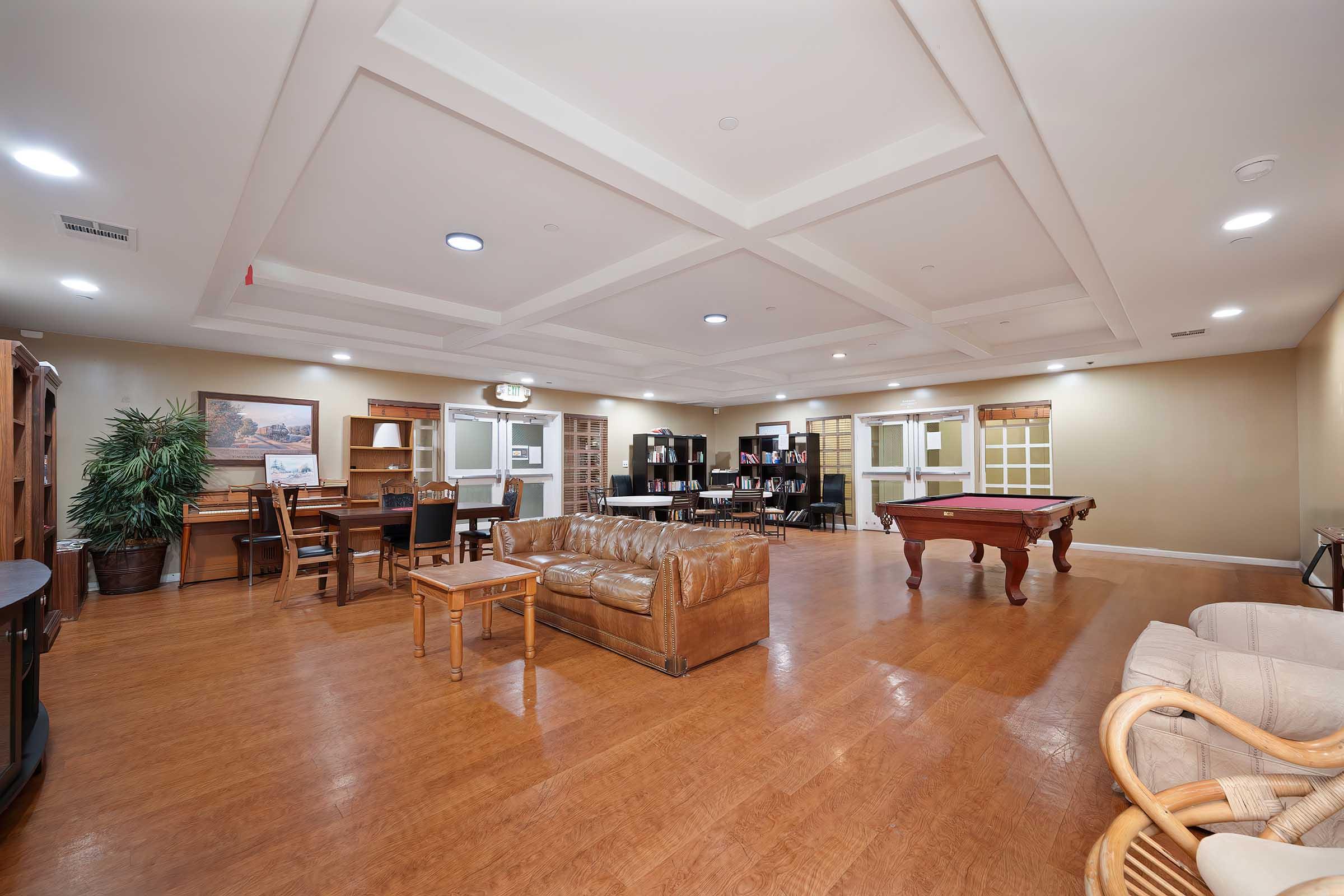 A spacious interior room featuring a brown leather couch, wooden furniture including a table and desk, and a pool table. The walls are light-colored with a coffered ceiling. There are several doors and a tall plant, creating an inviting and comfortable atmosphere.