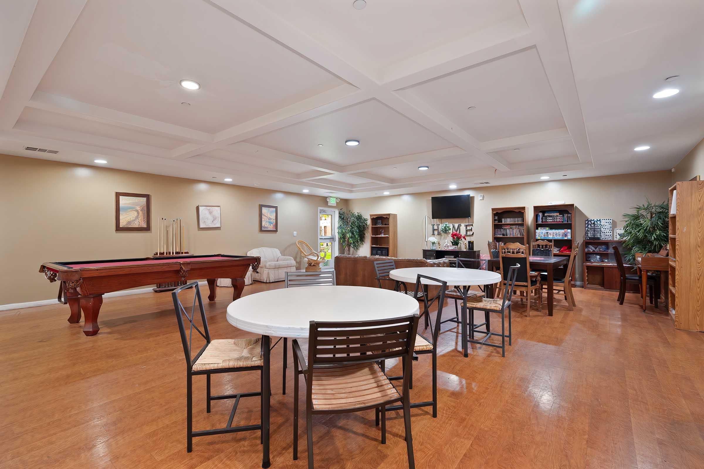 Interior view of a spacious recreation room with several round tables and chairs, a billiards table, a seating area, and entertainment options. The walls are painted in neutral tones, and there are decorative elements like framed pictures and plants. Large windows provide natural light in the space.