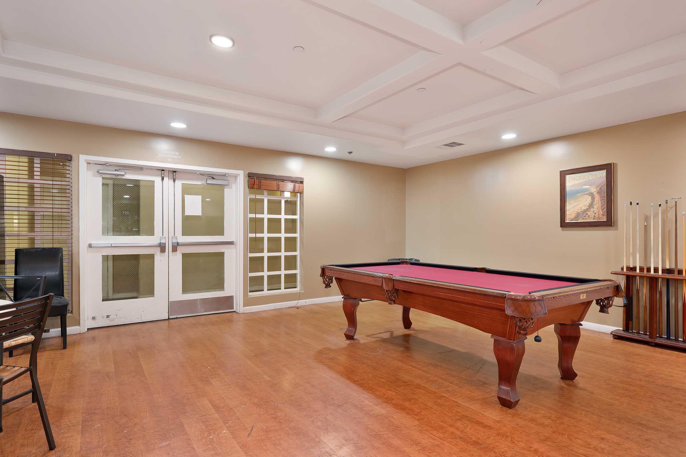 Interior view of a recreation room featuring a billiards table with a red felt surface, wooden flooring, and an artwork on the wall. There are glass doors leading to another space and a seating area with chairs. The ceiling has a grid pattern, and the room is well-lit with overhead lighting.