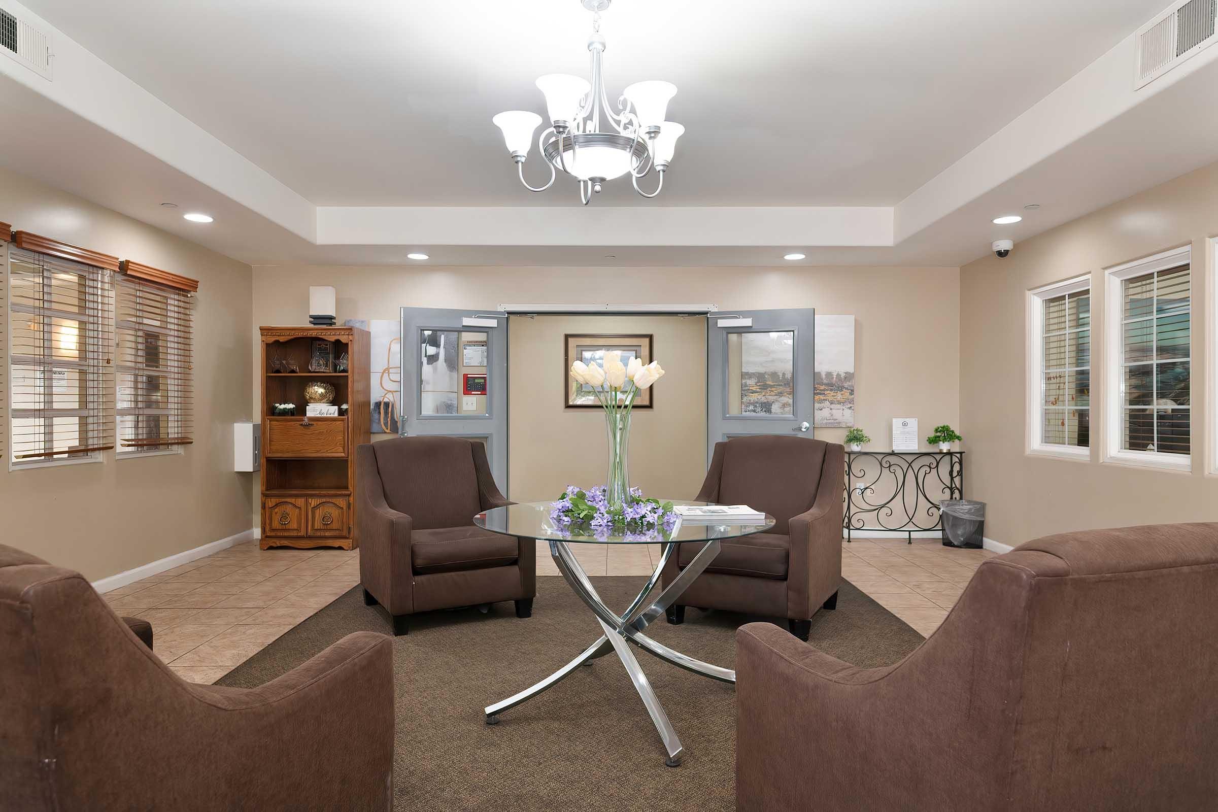 A cozy lobby area featuring two brown armchairs and a glass table with a vase of flowers. The space is well-lit with a chandelier and has beige walls, framed artwork, and windows allowing natural light. There are double doors leading to another area, alongside a wooden cabinet. The floor is tiled, adding to the inviting atmosphere.