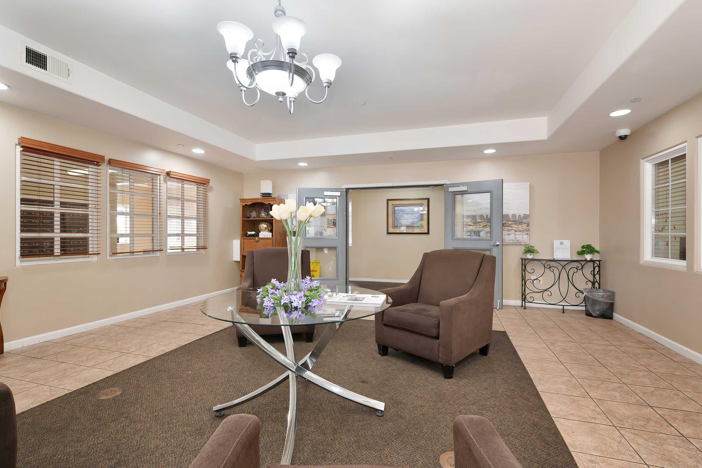 A cozy interior waiting area featuring two brown armchairs, a round glass table with a vase of white flowers, and a decorative bowl. Natural light streams through windows with wooden blinds, and a modern chandelier hangs from the ceiling. Wall art and a small shelving unit add to the welcoming decor.
