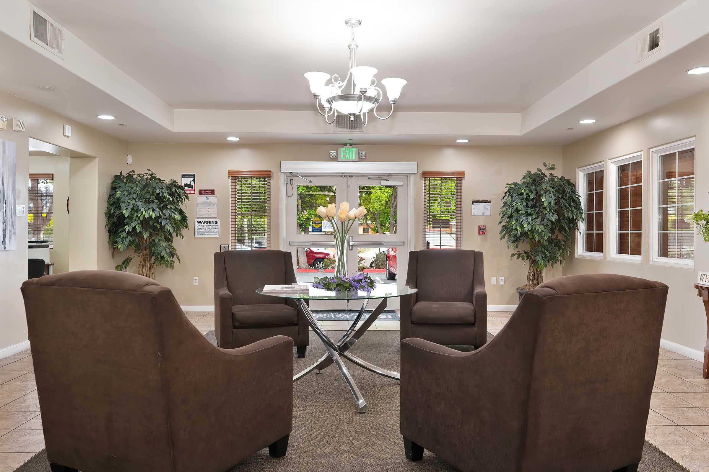 A welcoming lobby area with two brown armchairs facing each other, a glass-top coffee table in between, and a decorative arrangement of flowers on the table. The space features large windows with natural light, potted plants in the corners, and a chandelier overhead, creating a warm and inviting atmosphere.