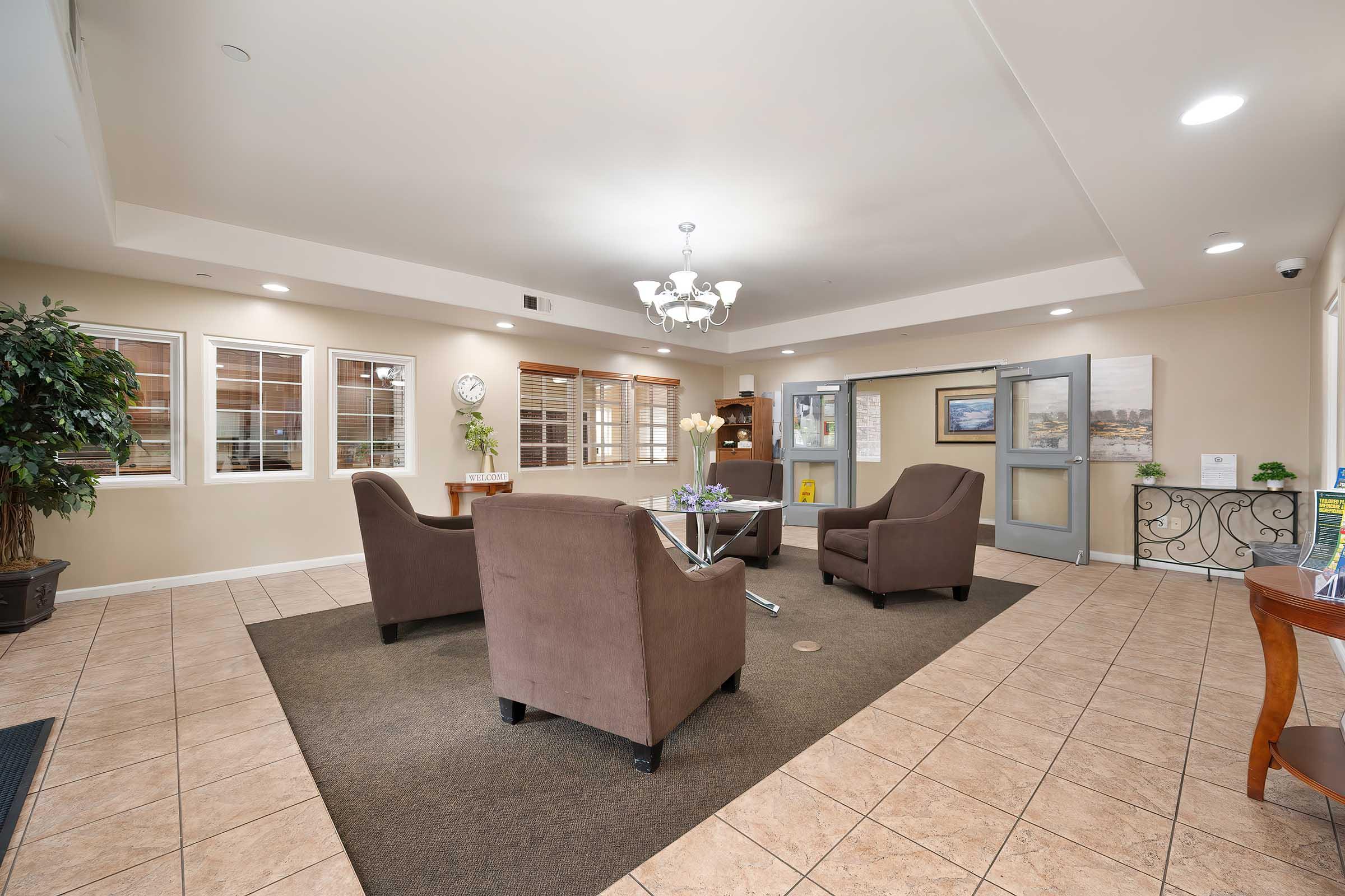 Interior view of a welcoming lobby area featuring four brown chairs arranged around a small table, a decorative plant in the corner, and large windows allowing natural light. The walls are painted a neutral color, and there is a chandelier overhead, creating a warm and inviting atmosphere.