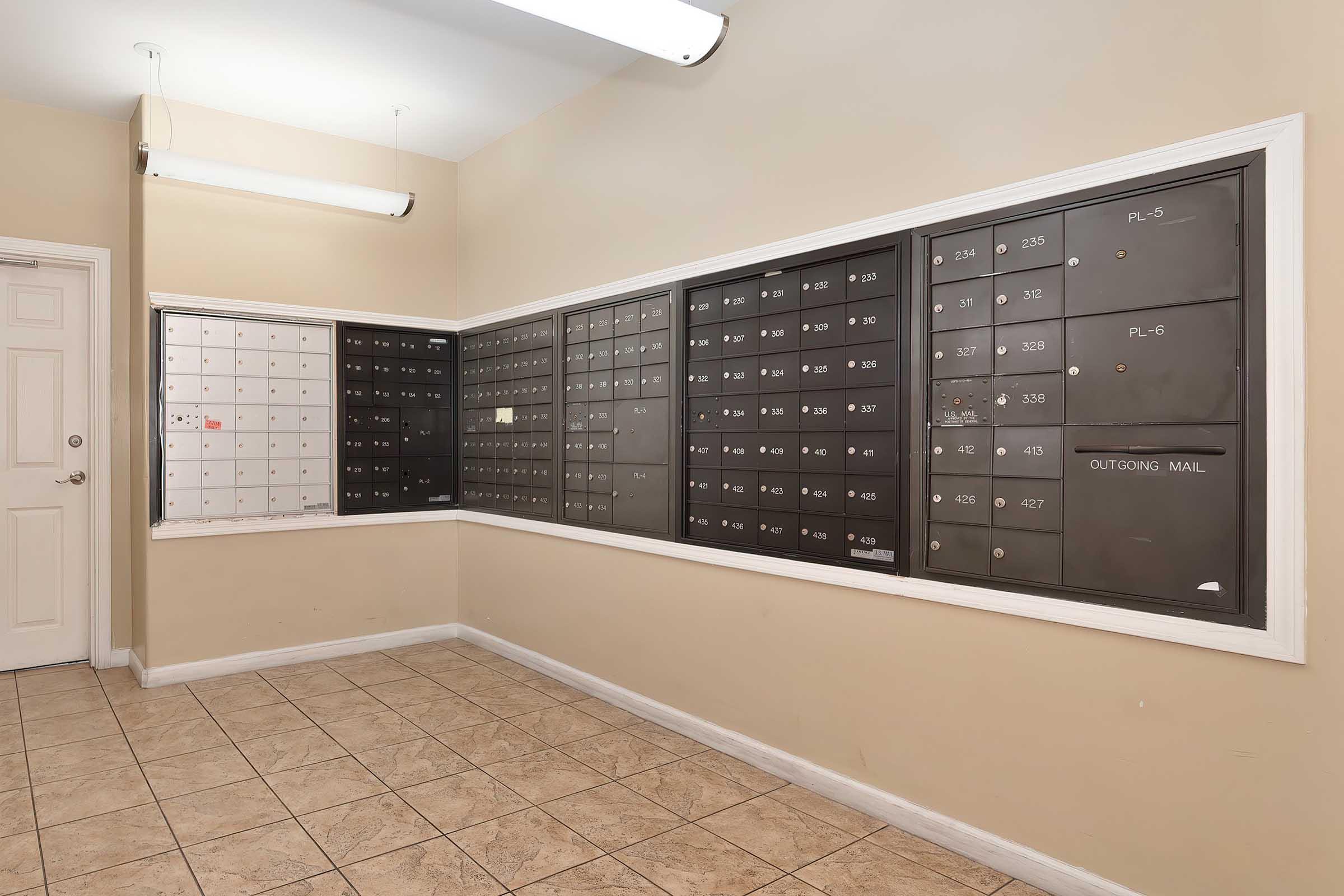 A mailroom with multiple wall-mounted mailboxes in two sections. The left section features a white mailbox panel with a few empty slots, while the right section contains dark-colored mailboxes with numbers, all set against a light beige wall and tiled floor. Ceiling lights illuminate the area.