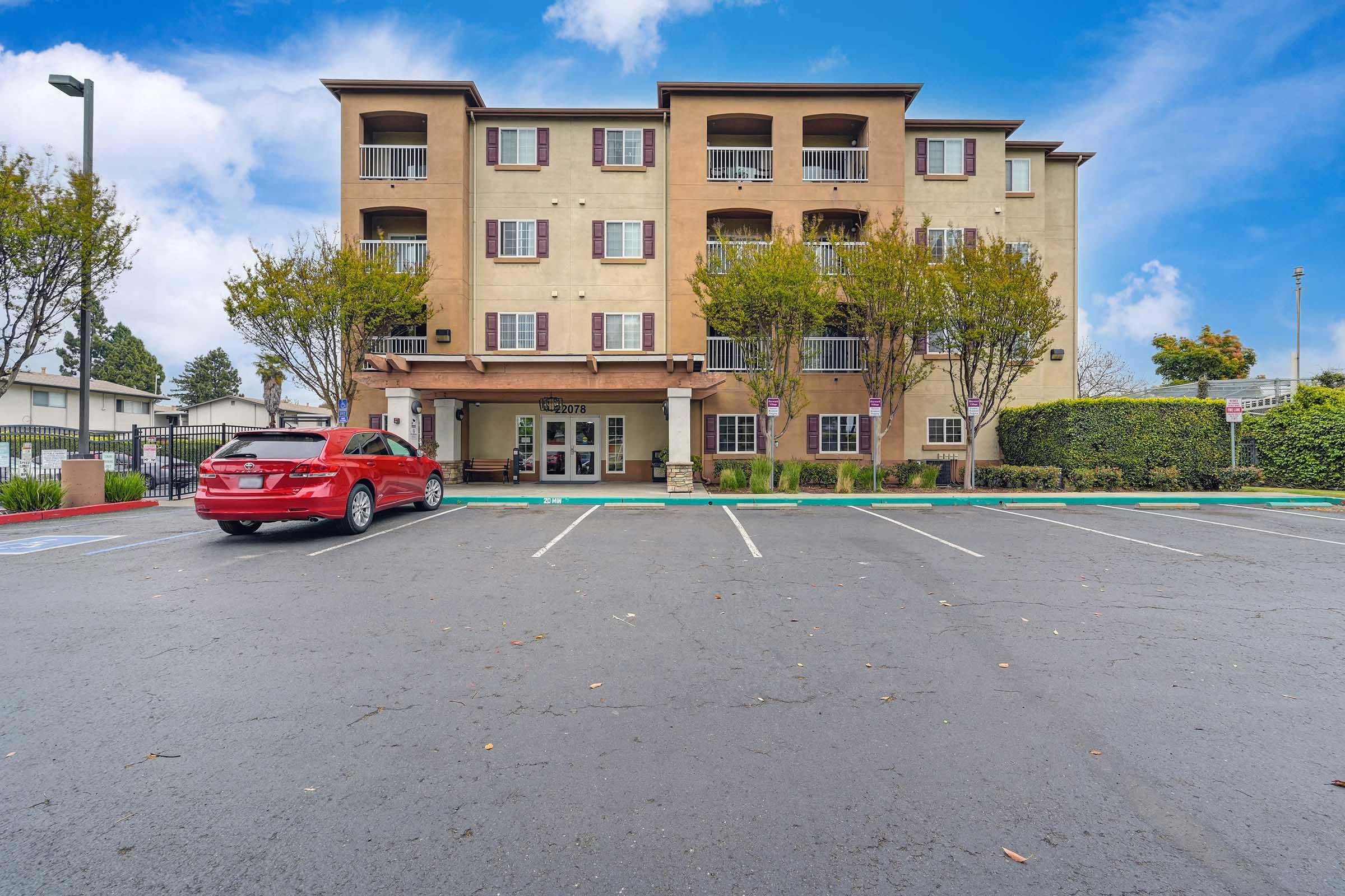 A four-story hotel building with brown exterior and multiple windows, surrounded by small trees and landscaping. A red car is parked in the foreground, and there are light blue skies with scattered clouds above. The hotel's entrance is visible, along with some parking spaces.