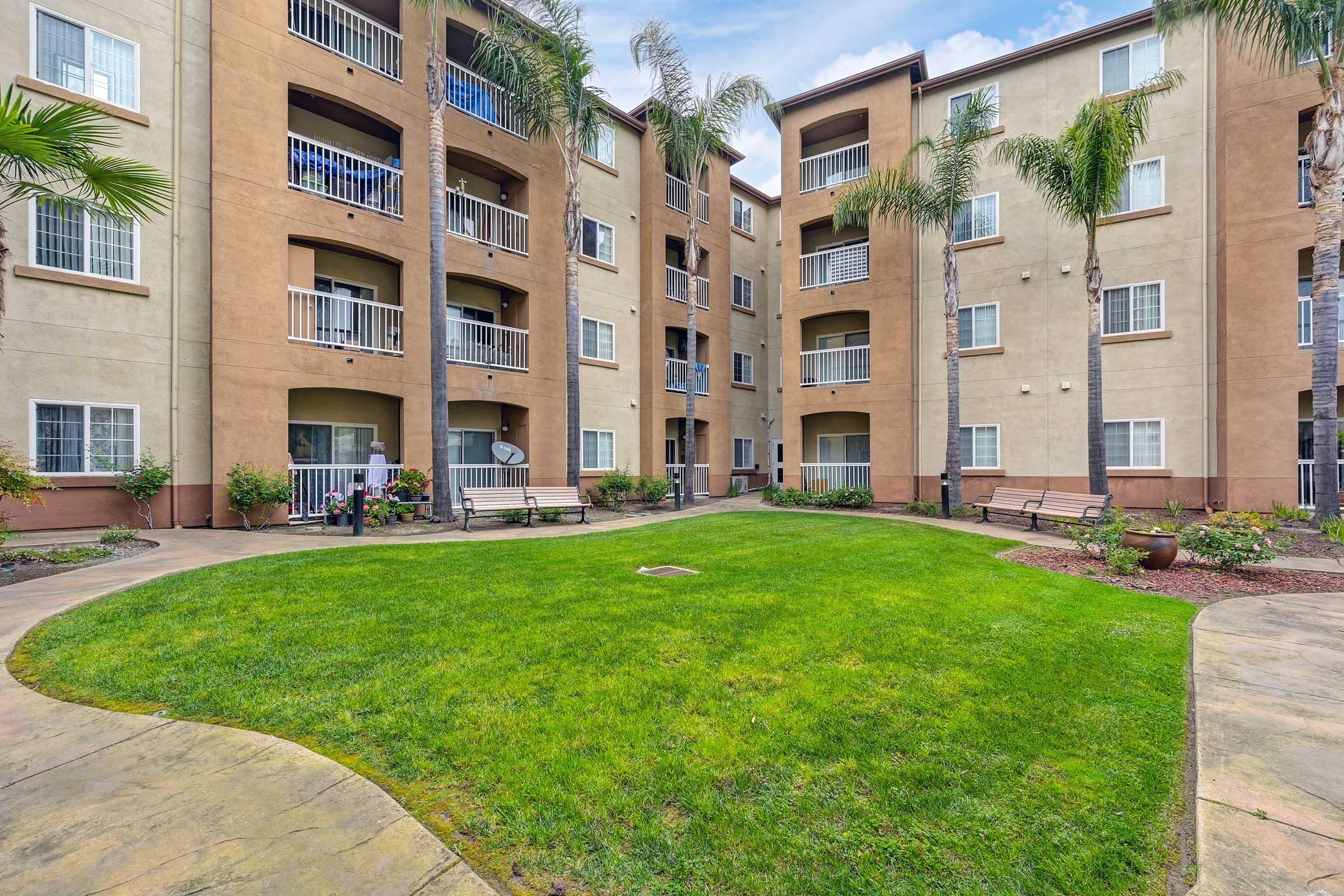 A well-maintained apartment complex courtyard featuring palm trees and neatly arranged benches. The grass is lush and green, providing a serene outdoor space. The buildings surrounding the courtyard are tan with multiple balconies, creating a welcoming residential environment.