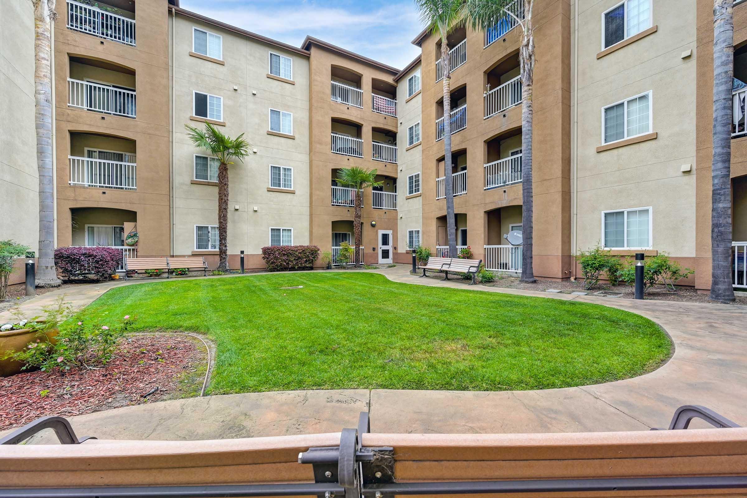 A landscaped courtyard of an apartment complex featuring green grass, palm trees, and benches. The buildings are three stories tall, with balconies on some units, and there are flowerbeds along the pathways. The image captures a peaceful outdoor area for residents to enjoy.