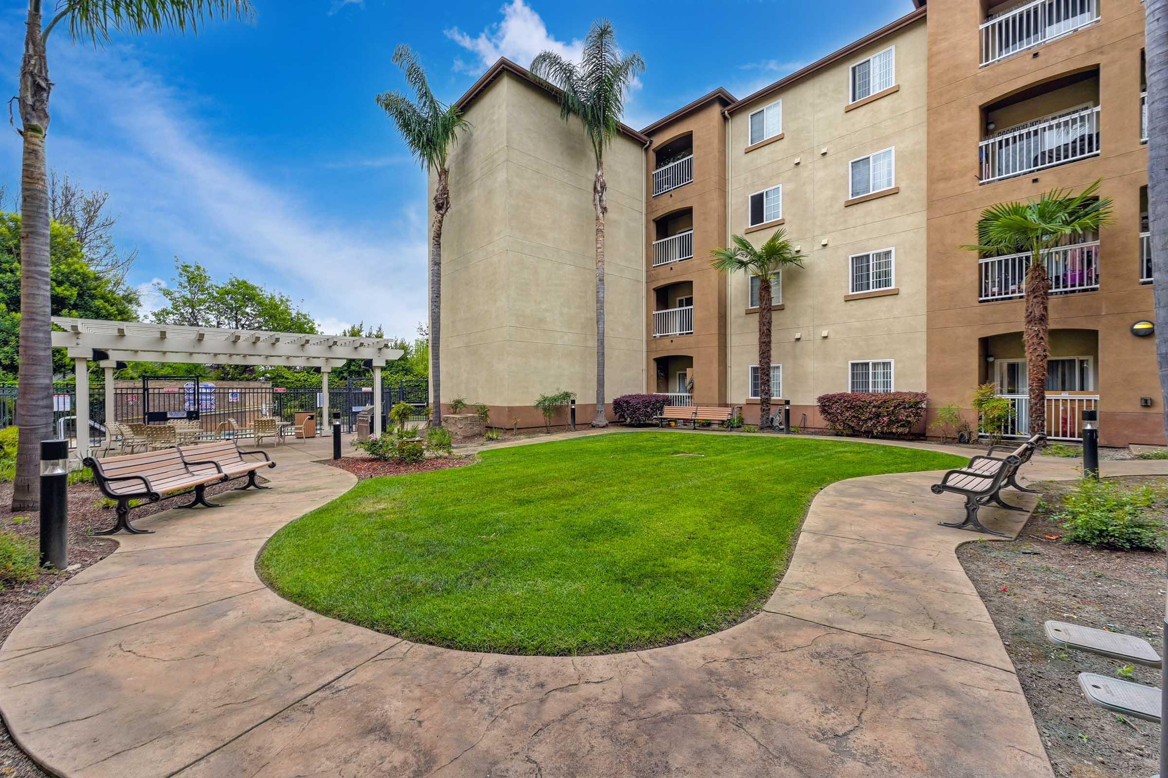 A well-maintained courtyard featuring lush green grass surrounded by palm trees. There are two benches for seating, a decorative pergola, and a multi-story apartment building in the background. Bright blue sky and greenery create a welcoming atmosphere.