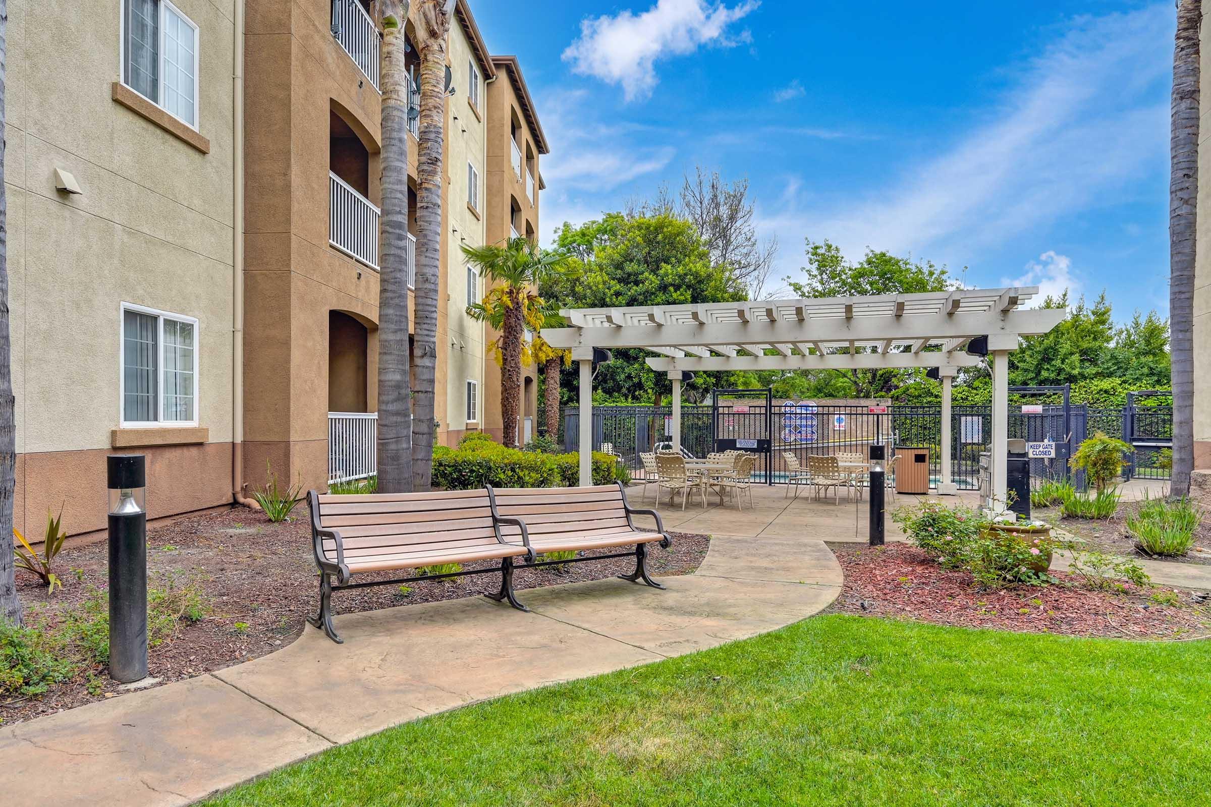 A landscaped area featuring a paved path leading to a shaded seating area with a pergola, surrounded by lush greenery and well-maintained flower beds. Nearby is an apartment building with a mix of stucco and stucco-like finishes, under a clear blue sky.