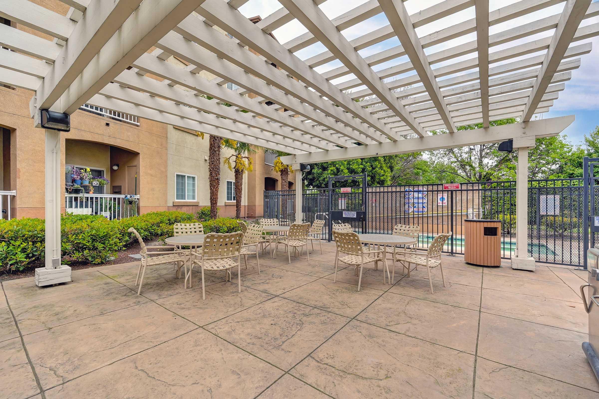 A shaded outdoor seating area with several chairs and tables beneath a white pergola, adjacent to a fenced swimming pool area. Lush greenery surrounds the space, creating a relaxing atmosphere for socializing or enjoying the outdoors. Clear blue skies are visible above.