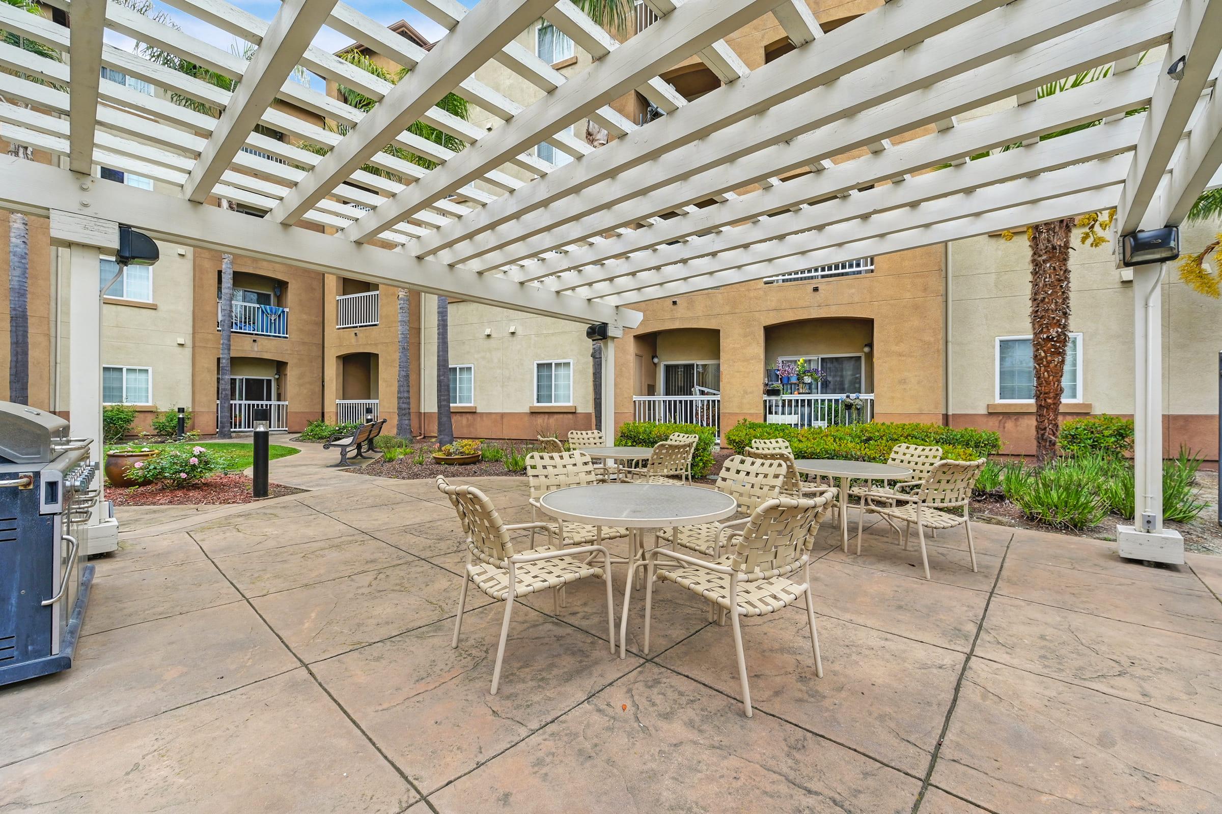 An outdoor patio area featuring a pergola with a white trellis overhead, surrounded by concrete flooring. There are several round tables with lightweight chairs, green landscaping and palm trees in the background, along with an outdoor grill to one side. Ideal for gatherings or relaxation.