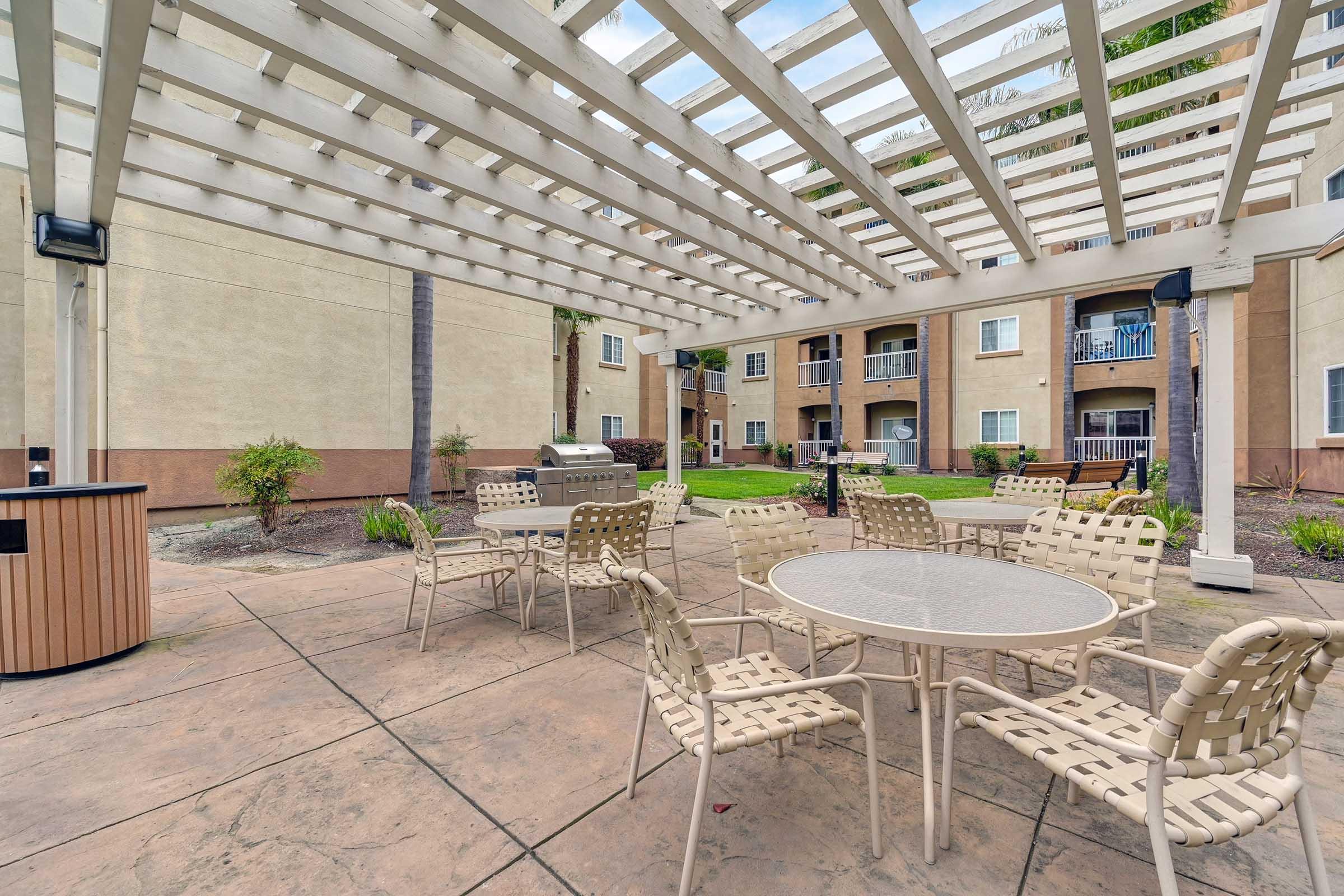 An outdoor seating area with a pergola, featuring several round tables and chairs. In the background, there are apartment buildings, and a barbecue area is visible. The ground is paved, and there are landscaping elements like plants and trees around the space. The sky is partly cloudy.