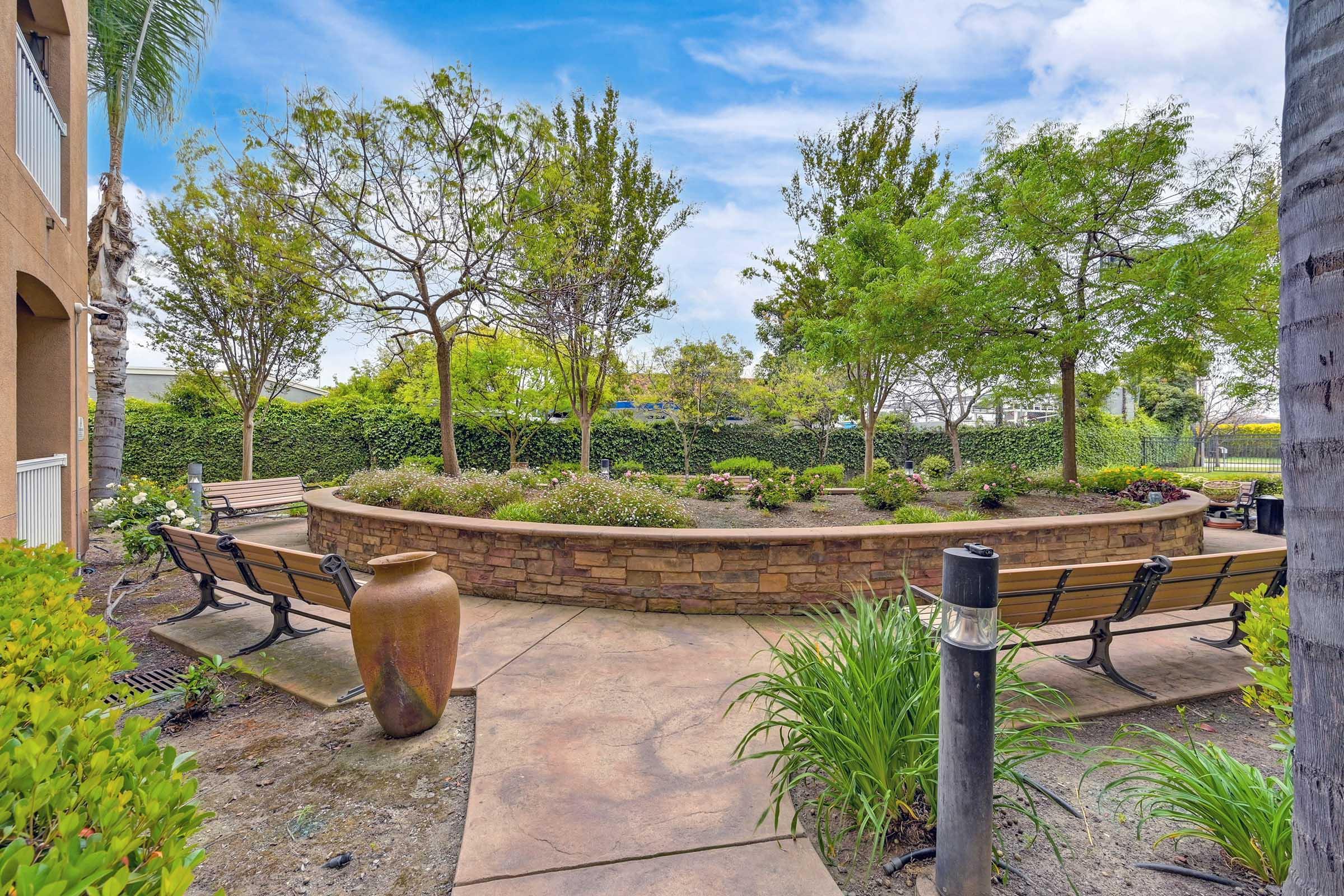 A serene outdoor space featuring a circular garden with benches and lush greenery. Trees and shrubs surround the area, creating a peaceful setting. A decorative pot is placed prominently, and the pathway is made of patterned stone, enhancing the inviting atmosphere of this landscaped environment.