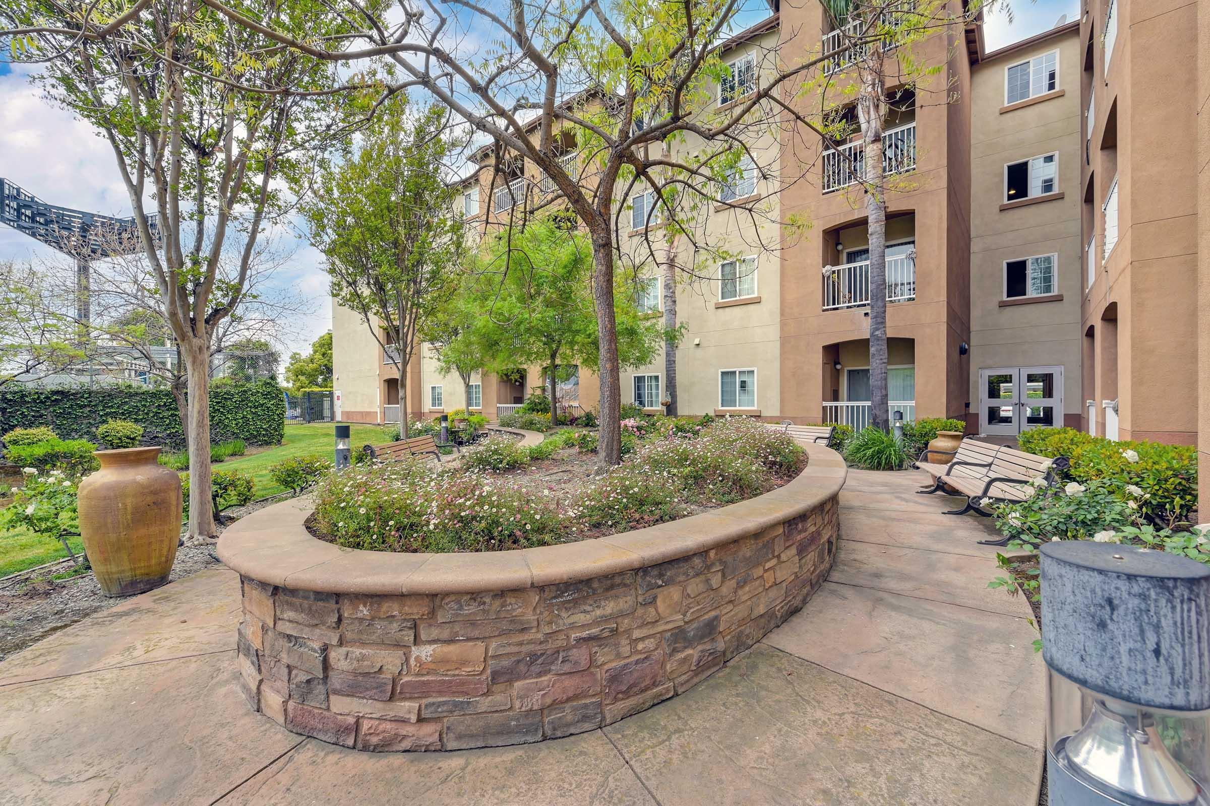 A landscaped courtyard featuring a stone-bordered garden with colorful flowers, surrounded by benches and trees. The background includes an apartment building with multiple balconies and windows, creating a peaceful outdoor space.