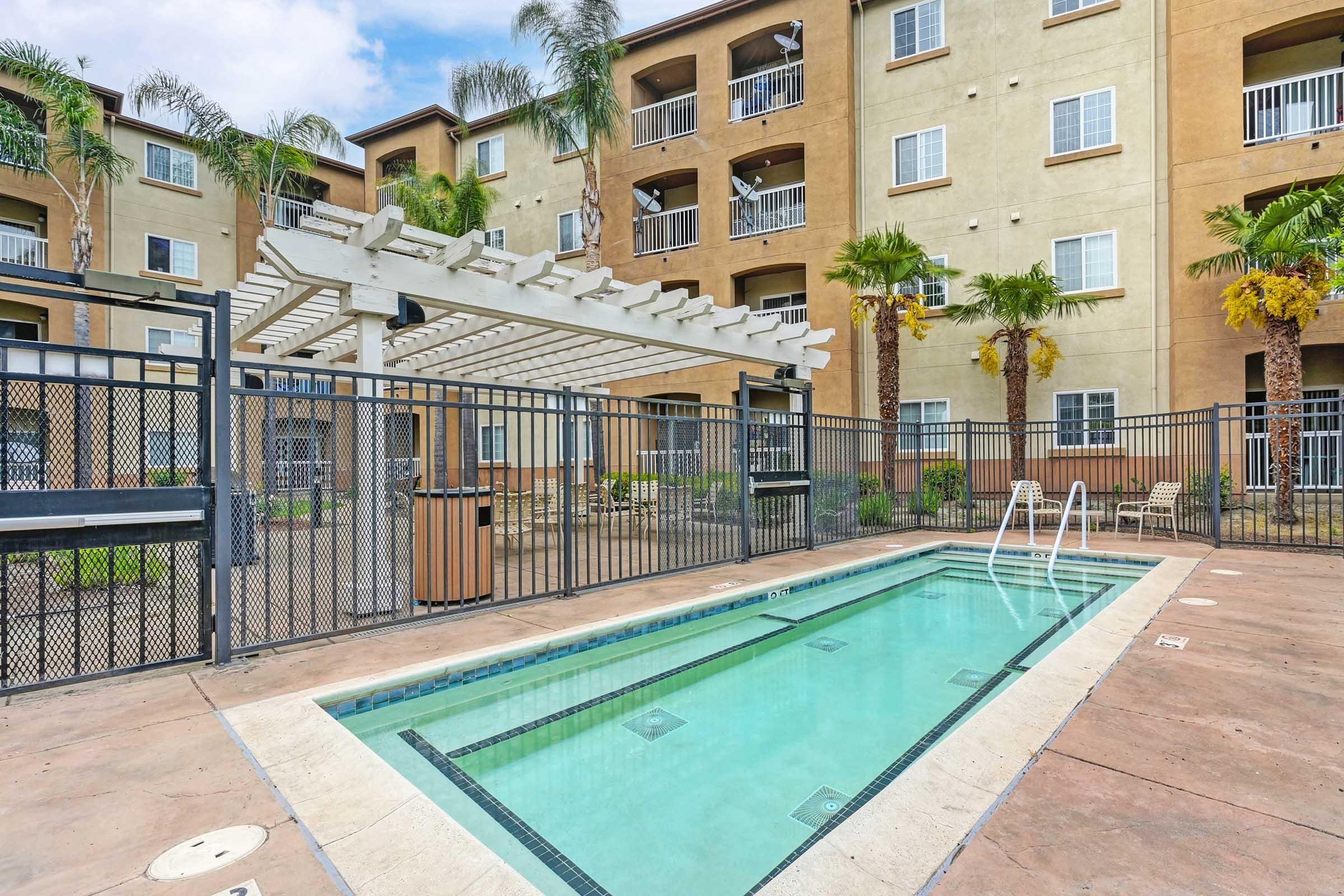 A swimming pool surrounded by a fenced area, featuring a white pergola and palm trees. The backdrop consists of a multi-story apartment building with balconies. The pool area has lounge spaces and is designed for relaxation in a residential setting.