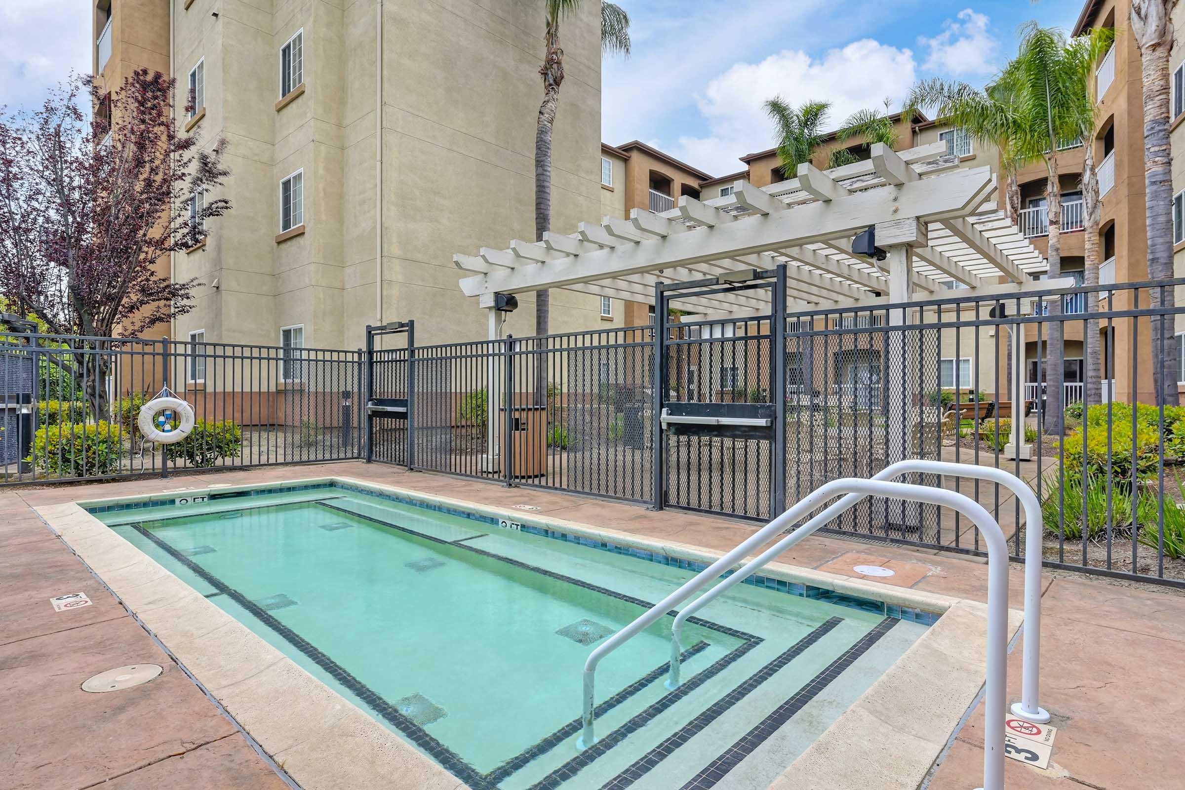 A small outdoor swimming pool is enclosed by a black fence, with a white pergola nearby. Lush greenery and palm trees are visible, and the backdrop features modern apartment buildings under a blue sky. The pool area includes safety features such as a lifebuoy.