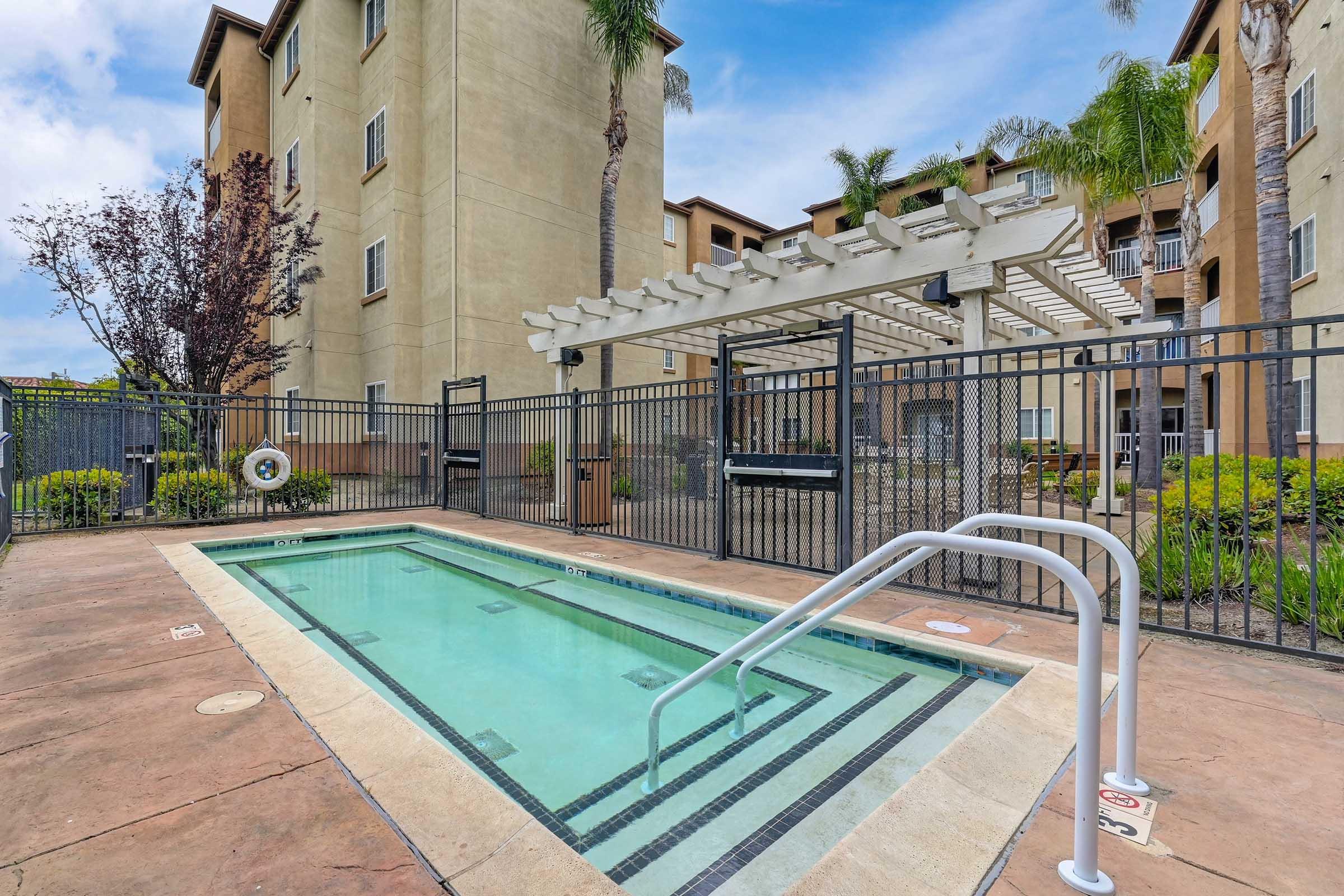 A gated outdoor pool area features a small swimming pool with a ladder, surrounded by concrete and landscaping. In the background, there are several multi-story buildings and a pergola with climbing plants. The sky is partly cloudy, creating a bright, inviting atmosphere.