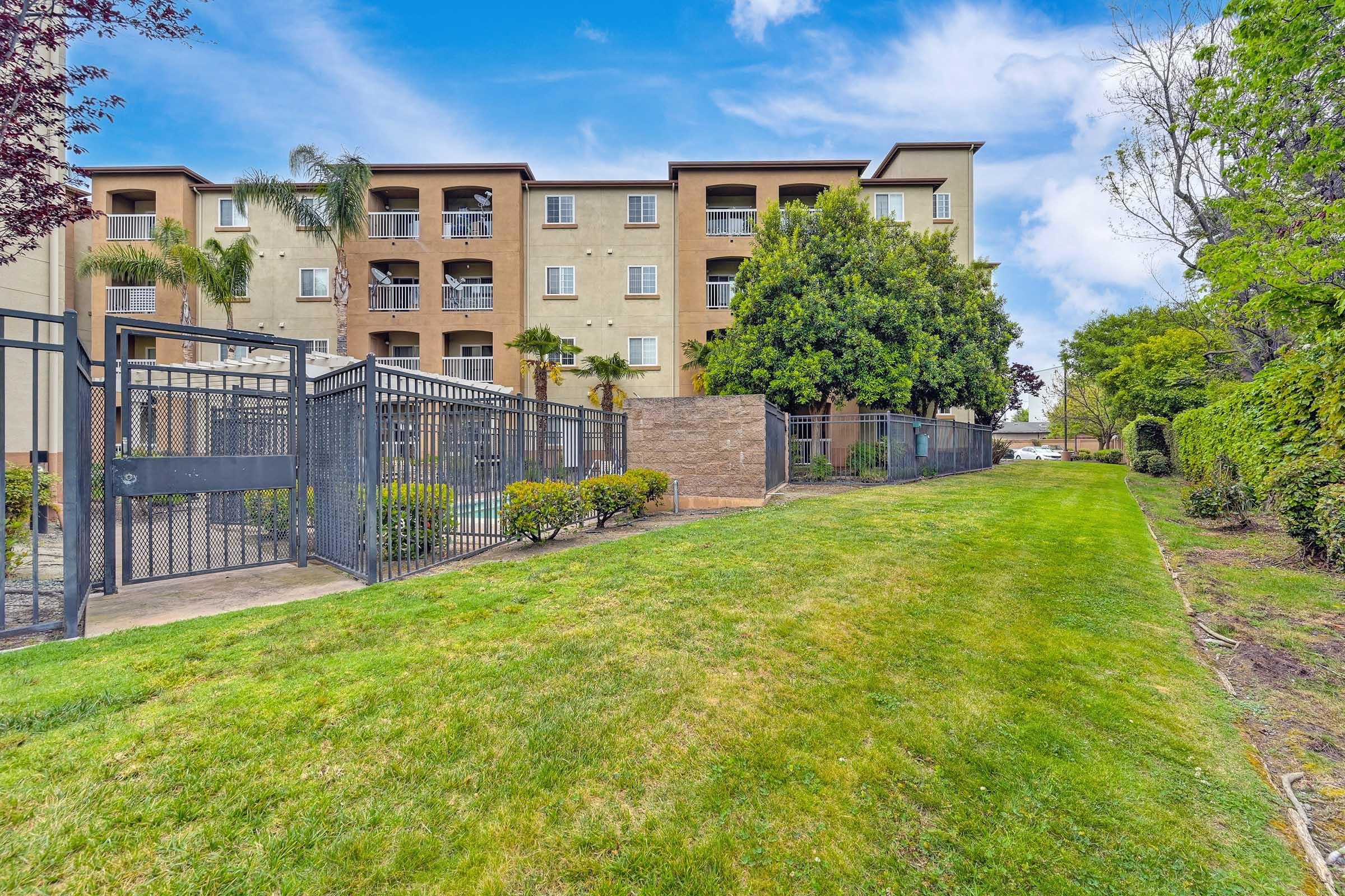 A well-maintained grassy area alongside a gated fence, leading to a multi-story apartment building with balconies and palm trees. The sky is bright and partly cloudy, creating a pleasant outdoor ambiance.